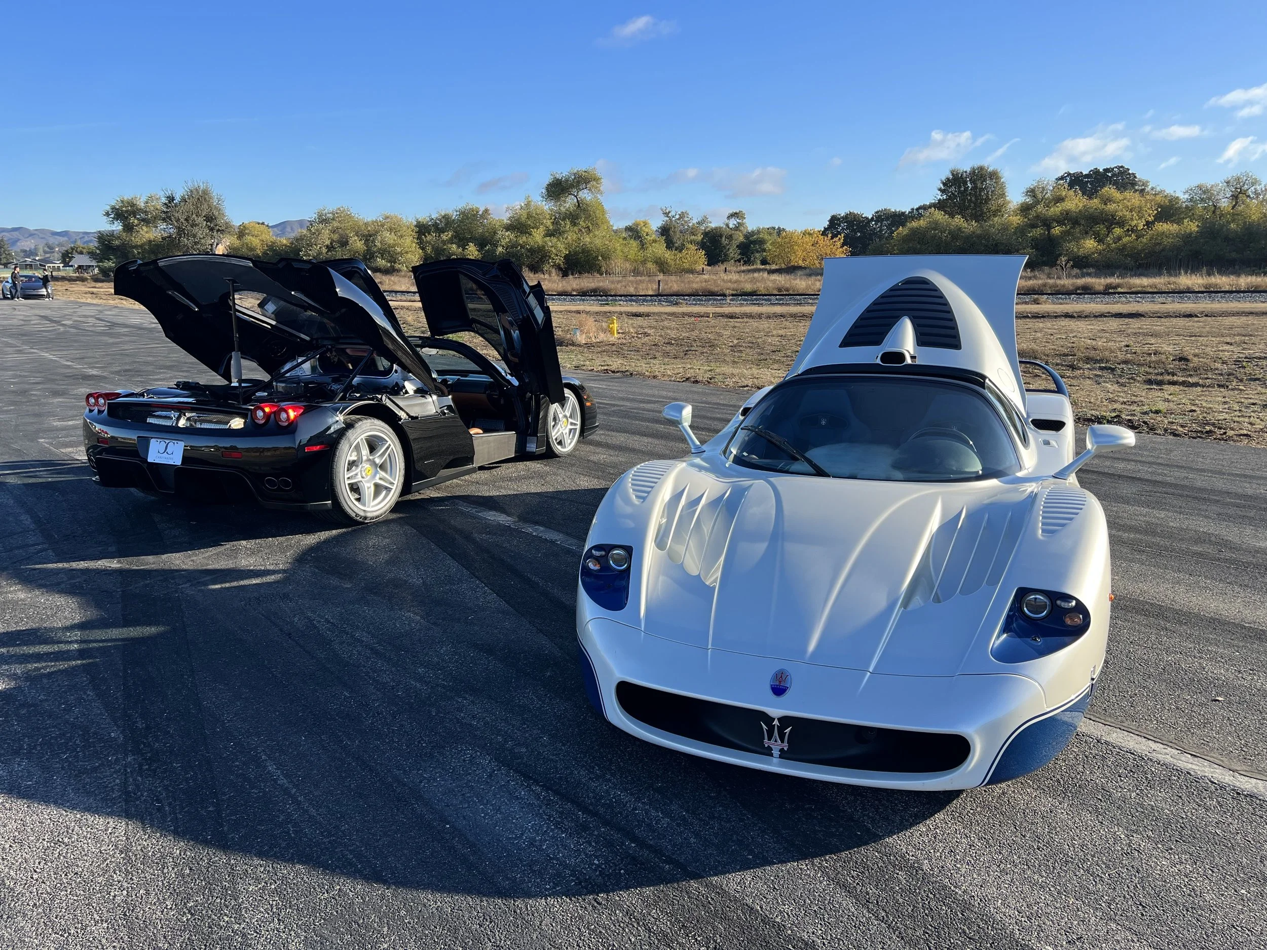 Two sports cars, a black Ferrari with its doors and trunk open and a white Maserati with its hood open, parked on an asphalt lot with a rural landscape in the background under a clear sky.