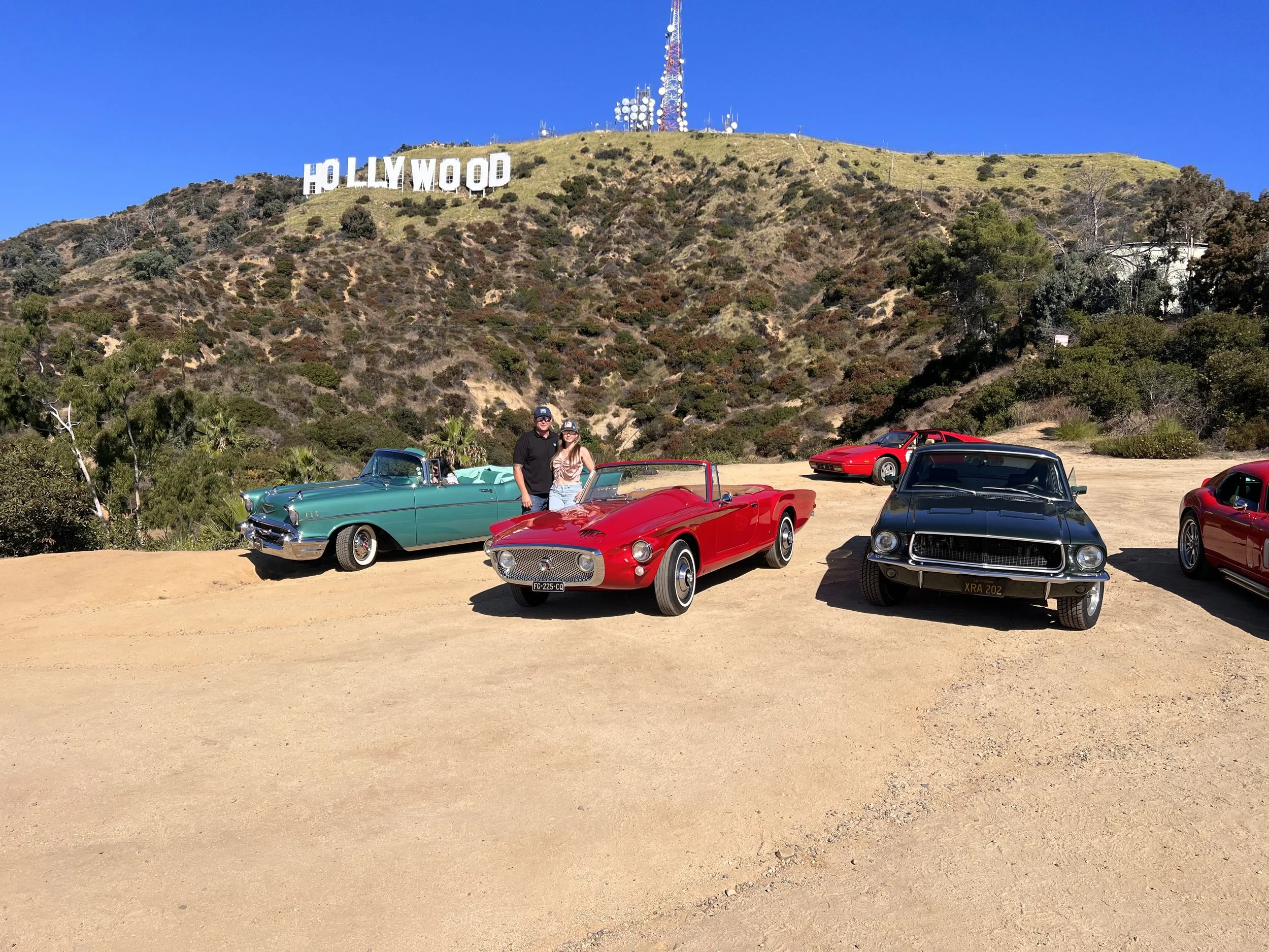 Vintage cars parked on a dirt lot with the Hollywood sign on a hillside in the background. Two people are standing near a teal classic car, and there are red and black cars nearby.
