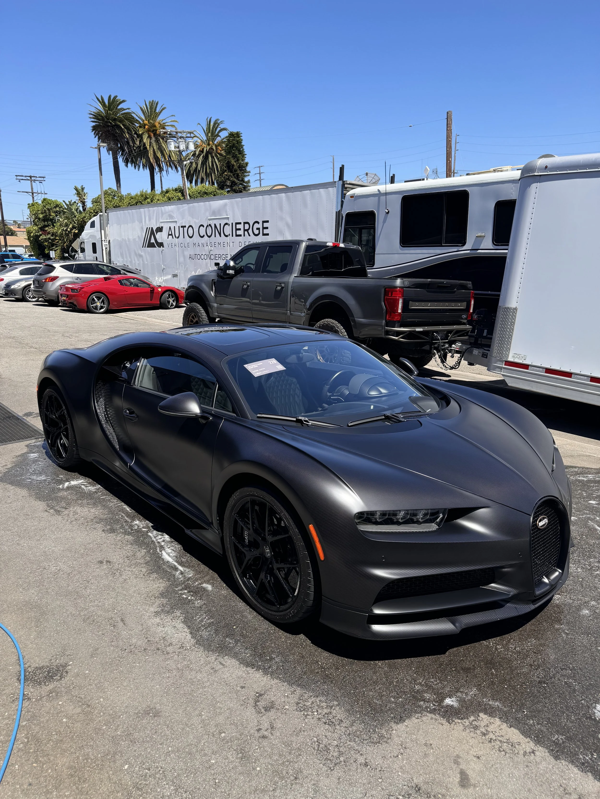A black Bugatti sports car parked outdoors in front of a trailer and other vehicles, including a black pickup truck and a red sports car, under a clear blue sky.