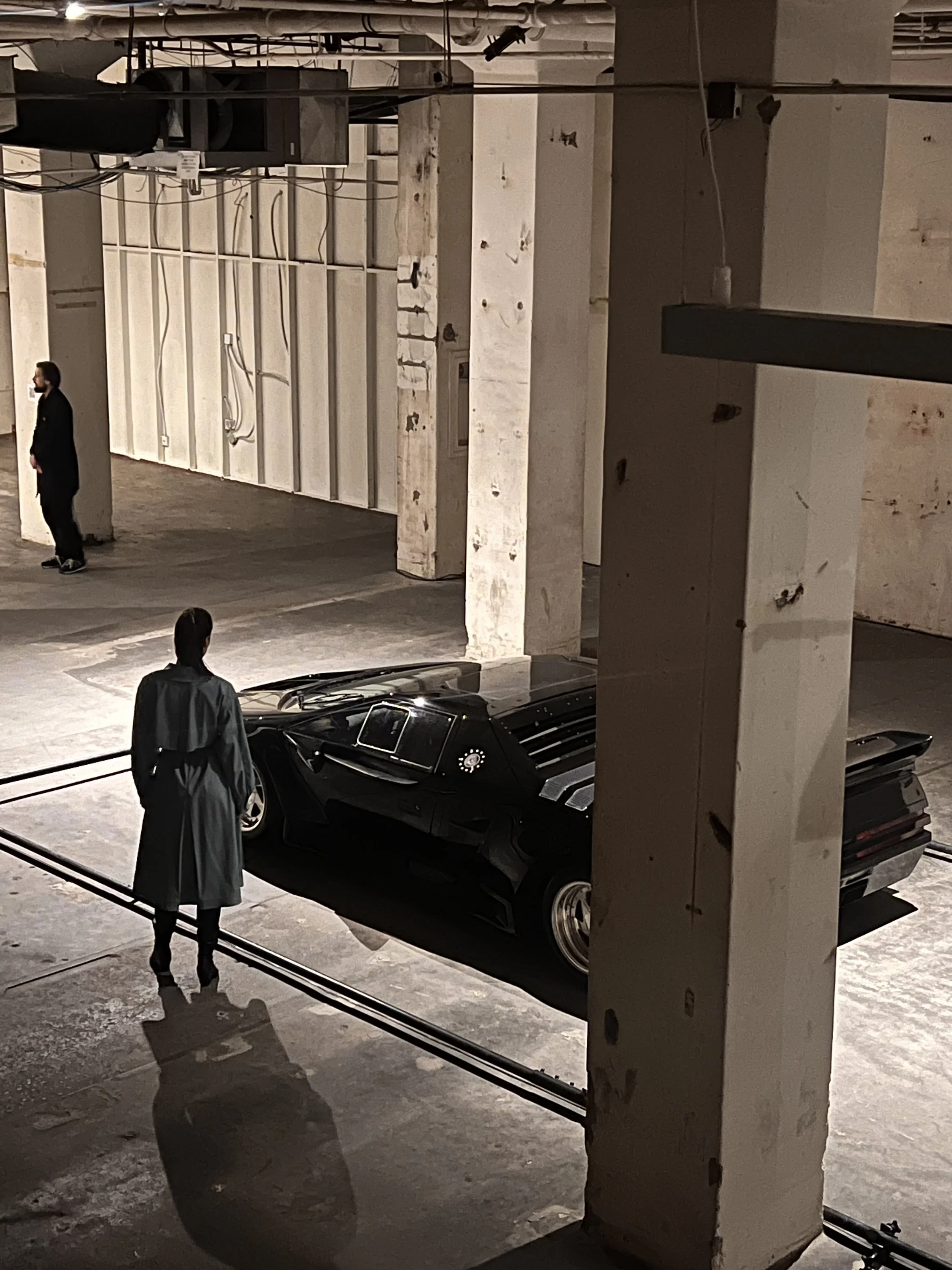 A woman stands in an industrial-style parking garage, looking at a black sports car parked near a concrete column.