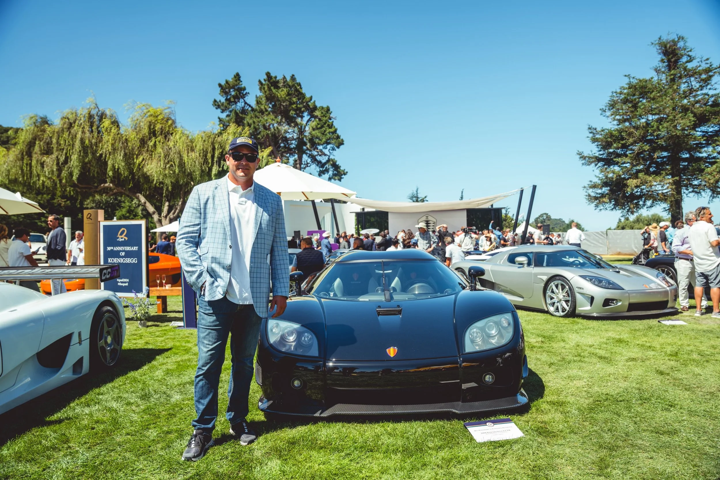 Man in a light blue checkered blazer and sunglasses standing next to a black sports car at a car show with other luxury cars and people in the background.