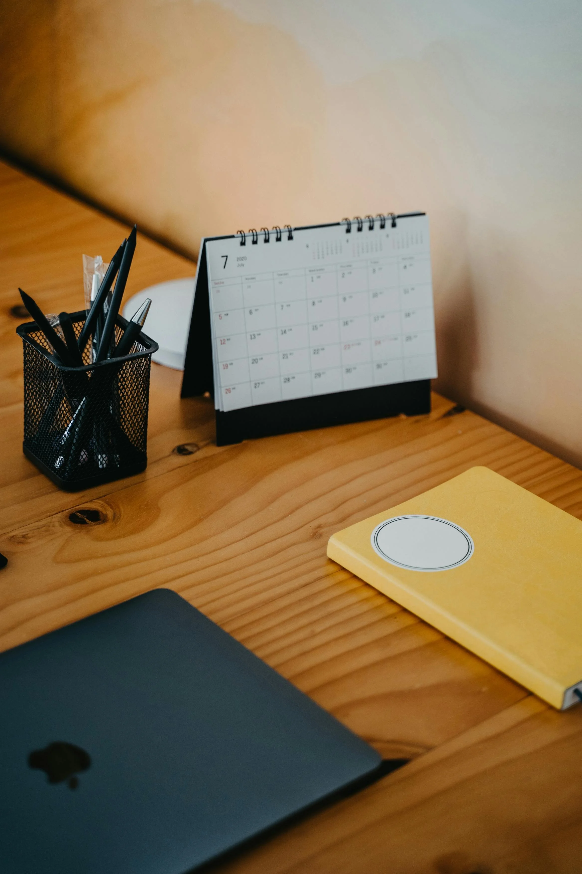 A wooden desk with a black pen holder containing black pens, a small calendar showing July 2020, a yellow notebook, a yellow sticky note, and part of a closed laptop.