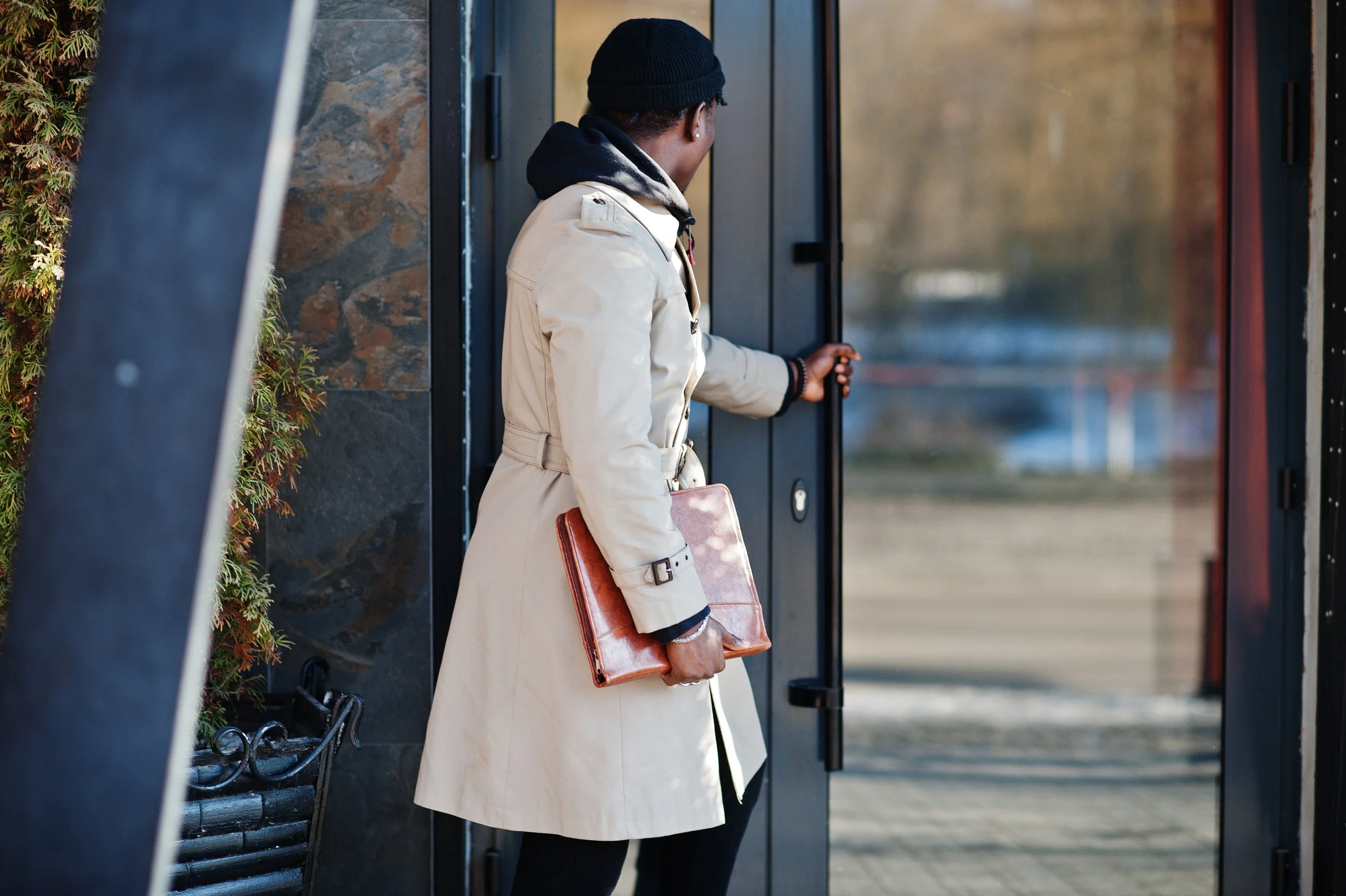A person wearing a beige trench coat, black beanie, and black hoodie is standing at a glass door, holding a brown leather bag, as they look inside.