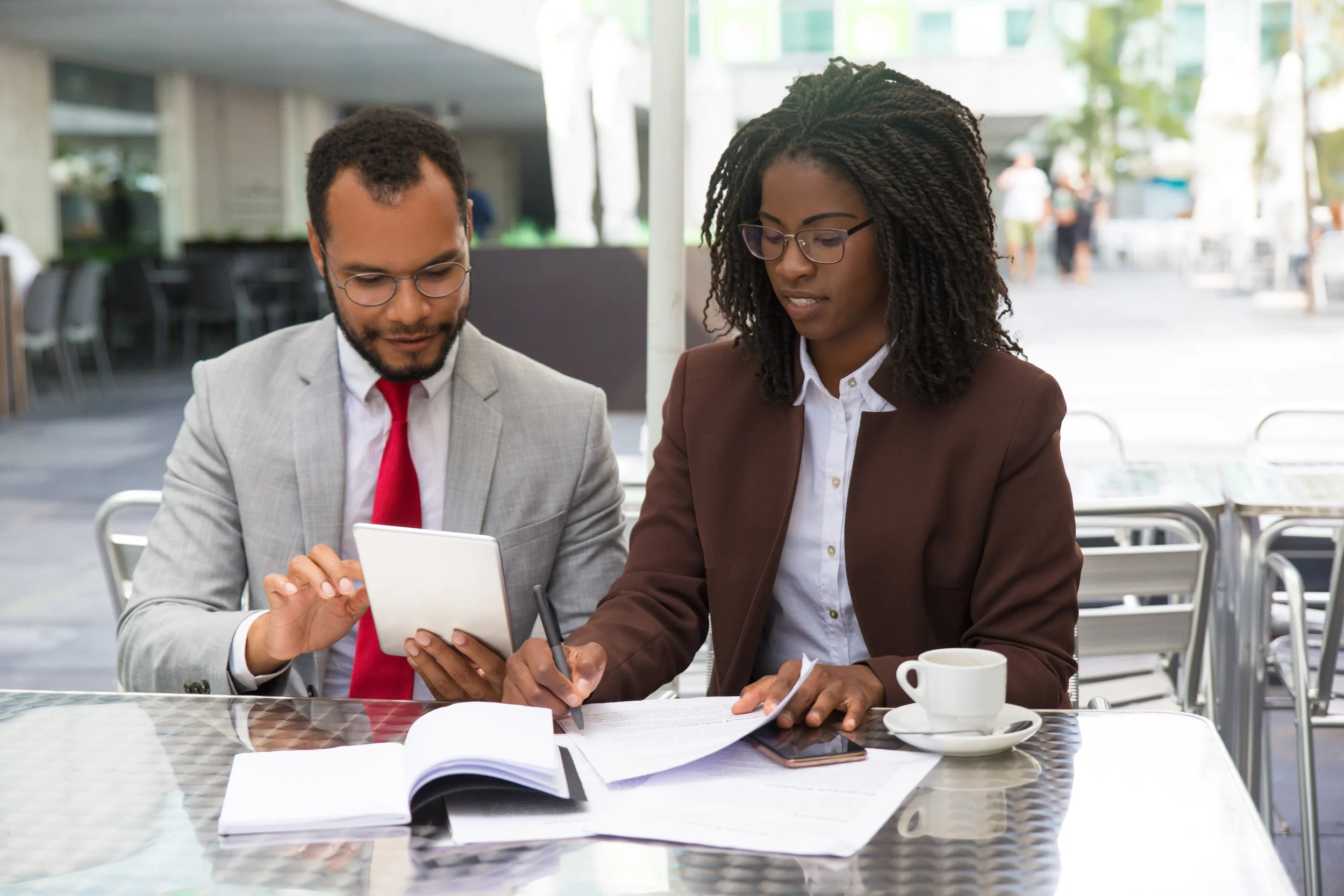 Two professionals, a man and a woman, sitting at an outdoor cafe table looking at a tablet. The man has a beard and wears glasses, a gray suit, and a red tie. The woman has curly hair, glasses, and wears a brown blazer and white blouse. The table has documents, a pen, a smartphone, and a white coffee cup.