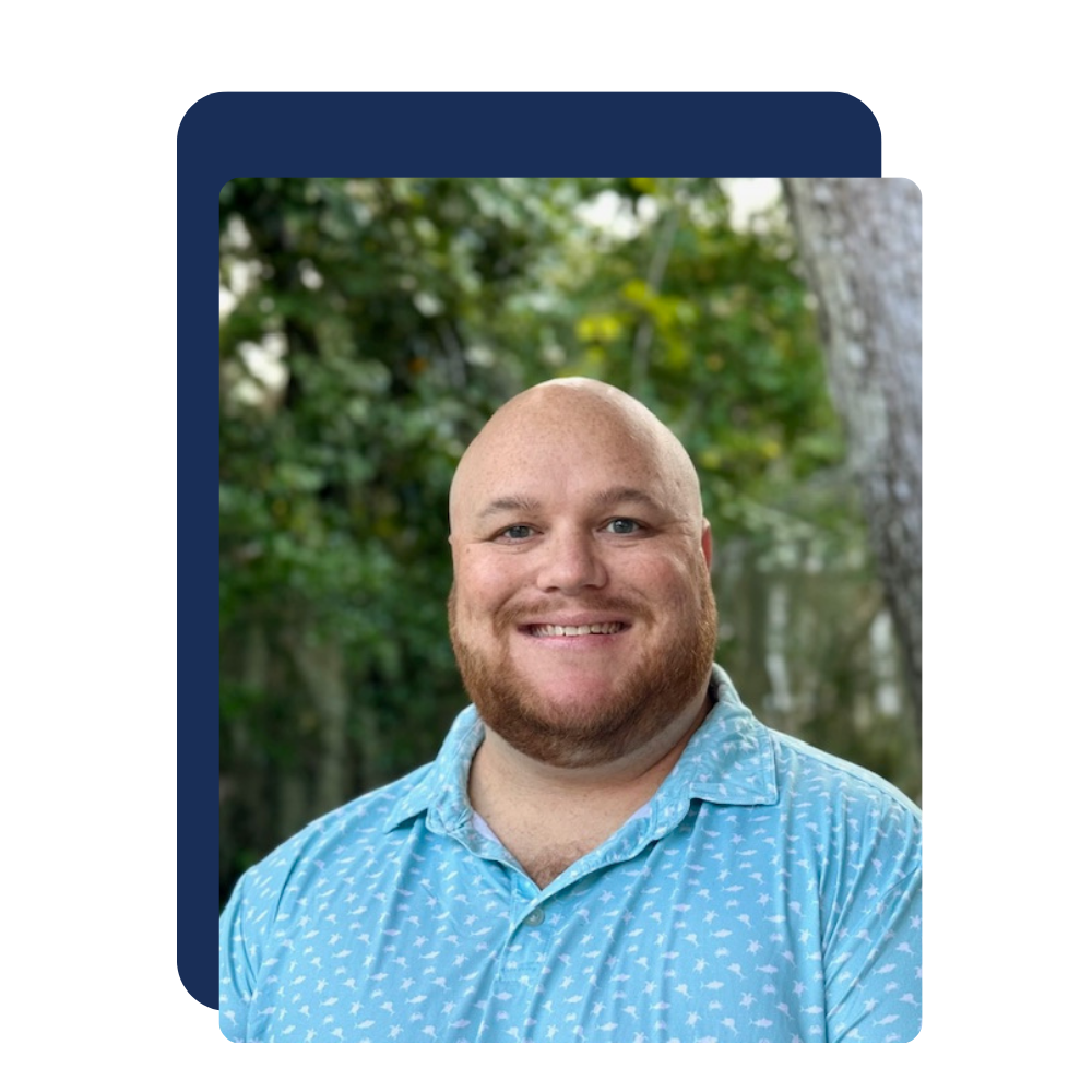 A smiling man with a shaved head and beard wearing a light blue, patterned button-up shirt, standing outdoors with a background of green trees and foliage.