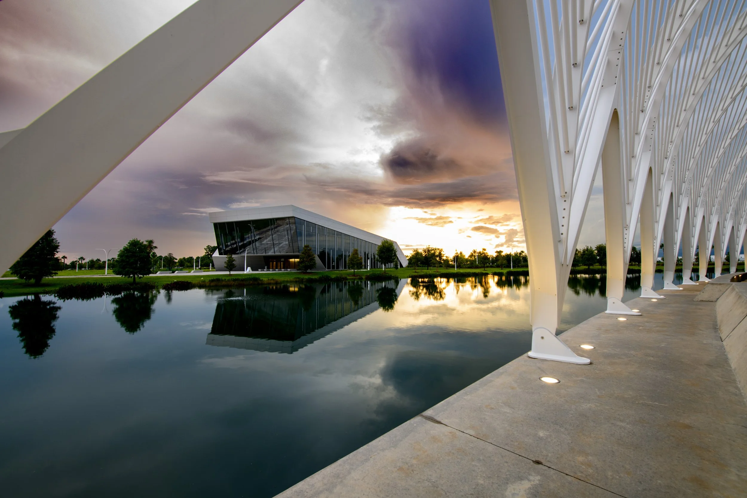 Modern building with glass facade across a water body, framed by white architectural structures at sunset with dramatic clouds and reflections in the water.