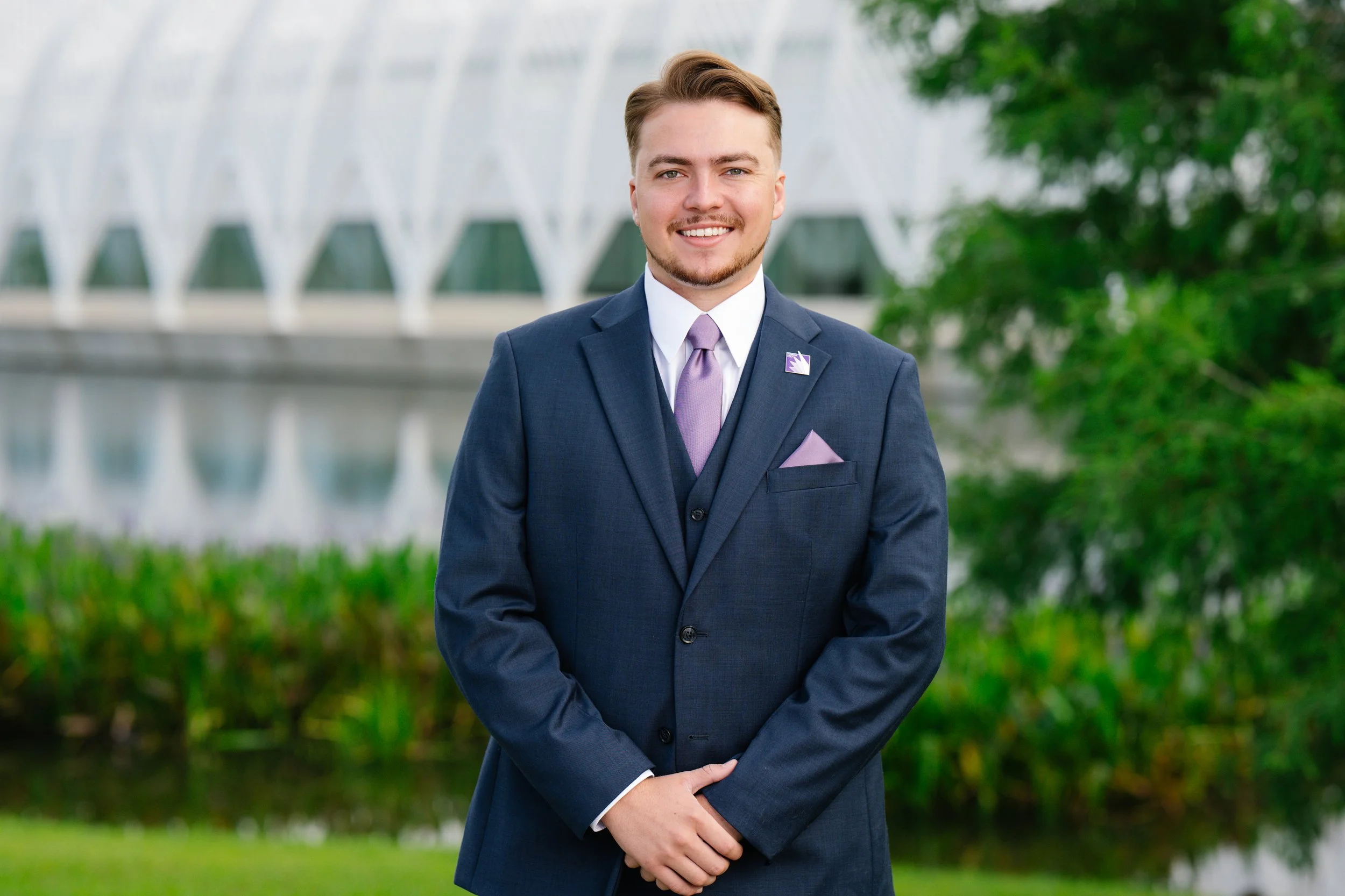 A young man in a dark blue suit and lavender tie standing outdoors near a modern building and greenery.