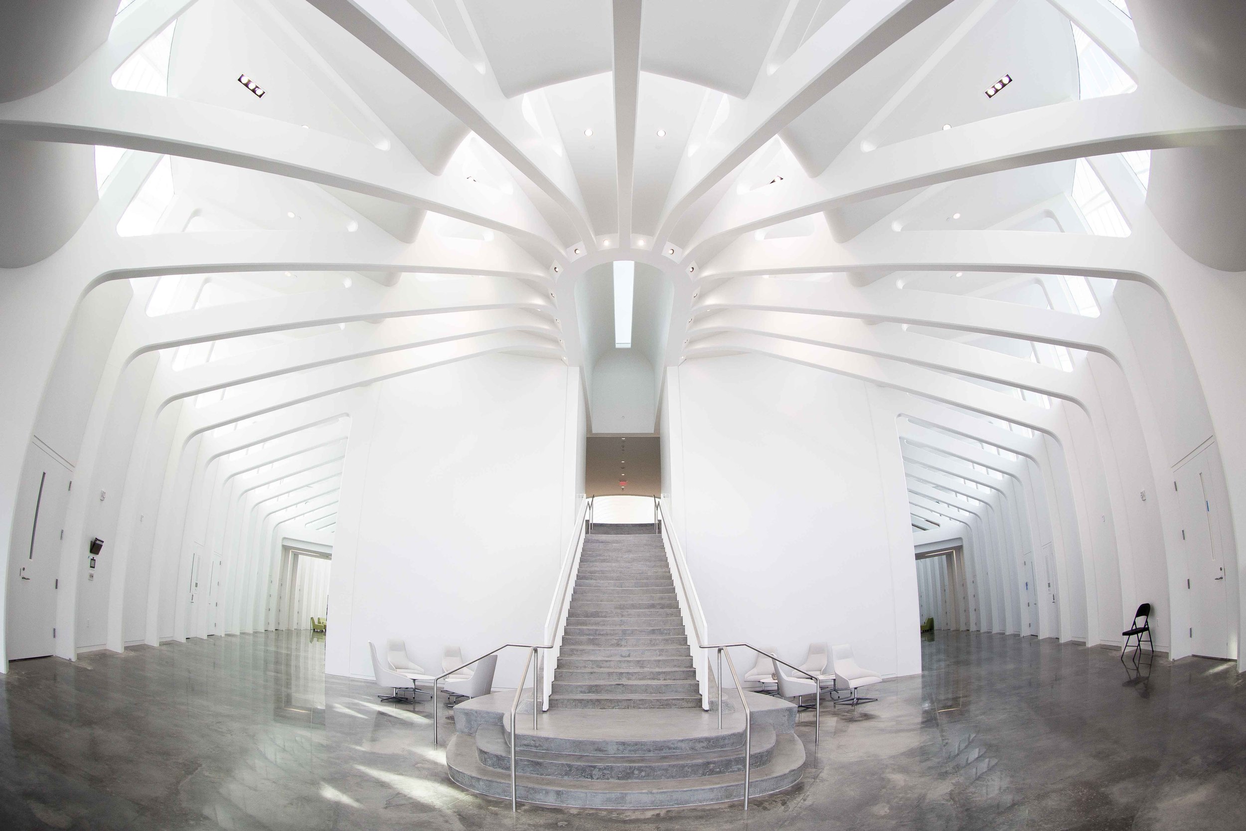 Modern white interior with a staircase in the center leading to an upper level, surrounded by symmetrical curved architectural elements and large windows that let in natural light, with seating on both sides.