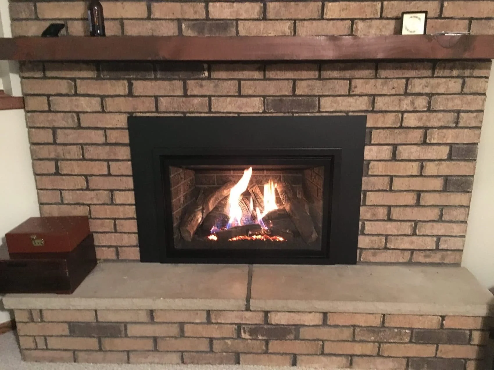 A brick fireplace with a black metal frame containing a lit fire. There is a wooden mantel on top with a few small items including a bottle and a small framed picture. On the left side of the fireplace, there is a small wooden box with a brass latch.