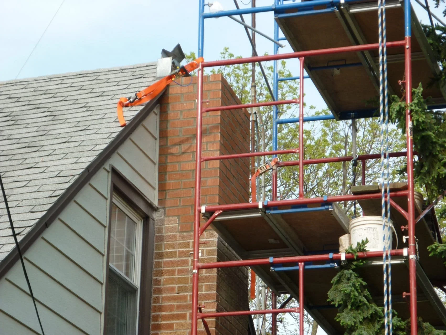 A house with a brick chimney and an adjacent section with gray wooden siding. A window with white trim is visible below the roof. Red and blue scaffolding is set up along the side of the house, with wooden planks on the scaffolding and a bucket at th