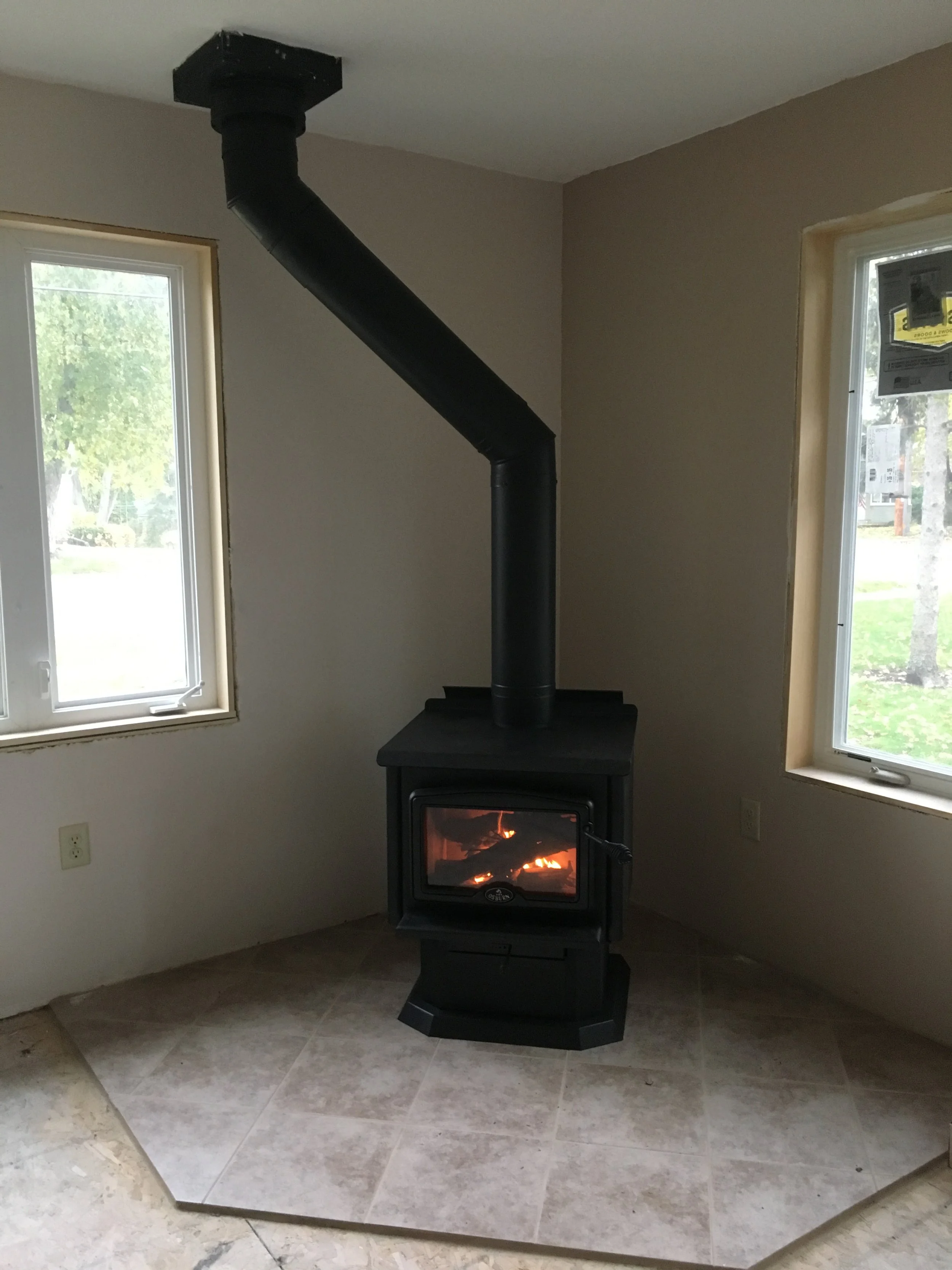 A black wood stove with a glass door burning logs, situated in a room with beige tiled floor and two windows on adjacent walls.