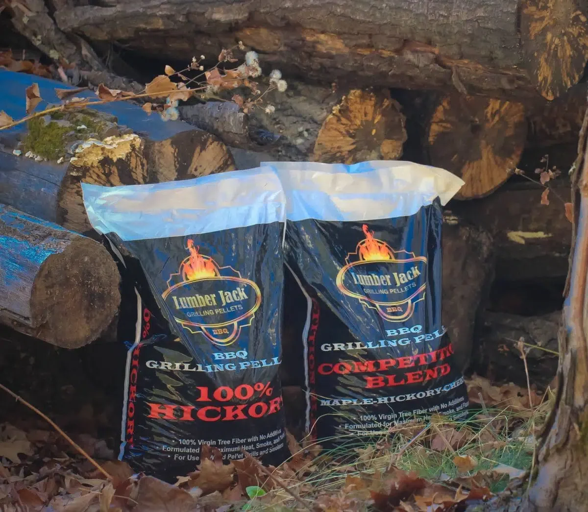Two bags of Lumber Jack BBQ grilling pellets, labeled as 100% Hickory and maple-hickory blended, positioned outdoors among logs and fallen leaves.