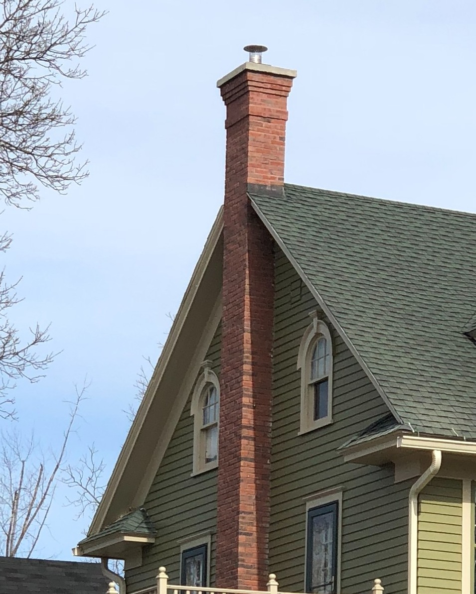 Close-up of the upper portion of a house showing a brick chimney and green gabled roof with two arched windows.