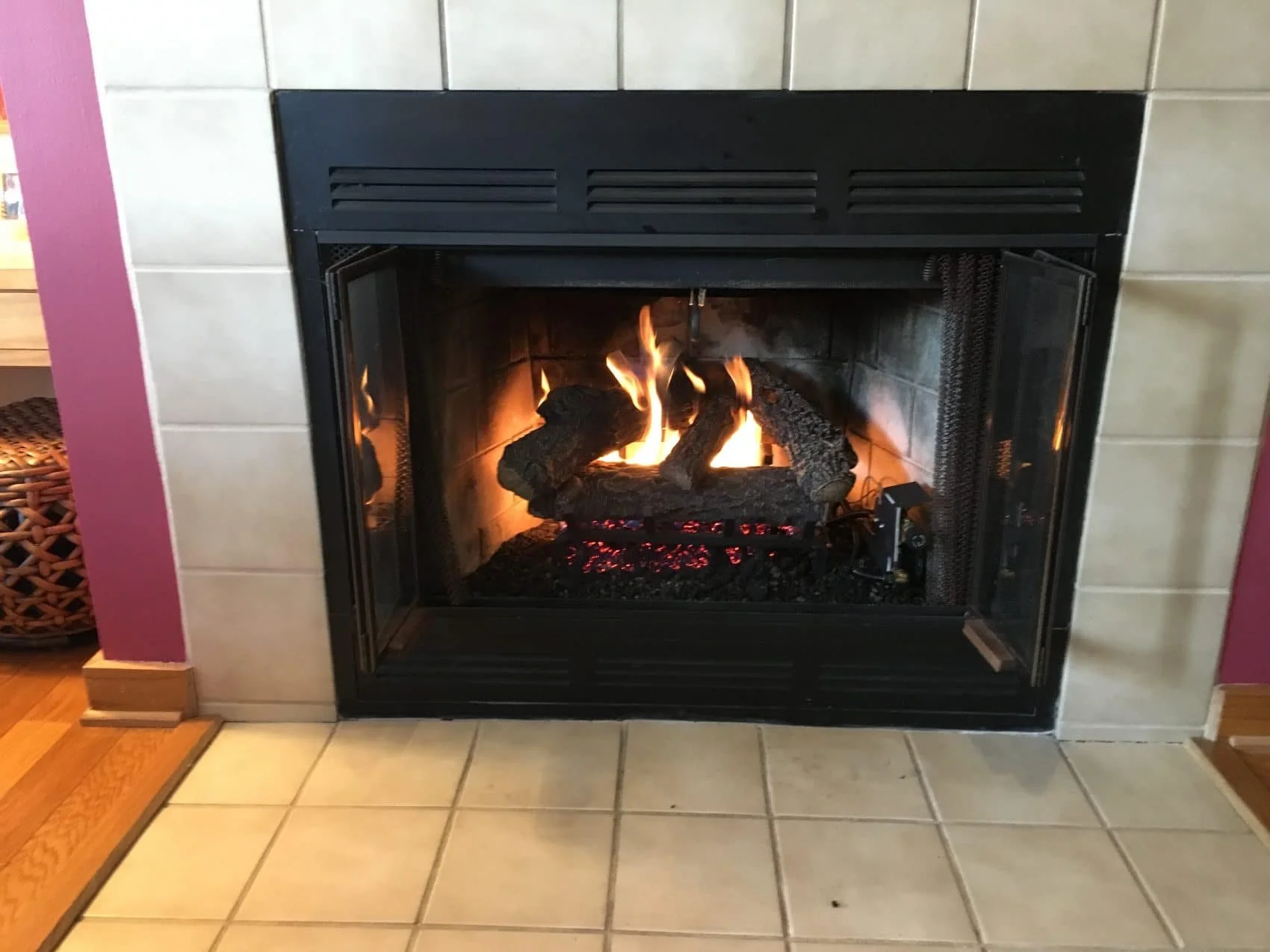 A lit gas fireplace with burning logs inside a room, with tiles around it and a purple wall to the left.