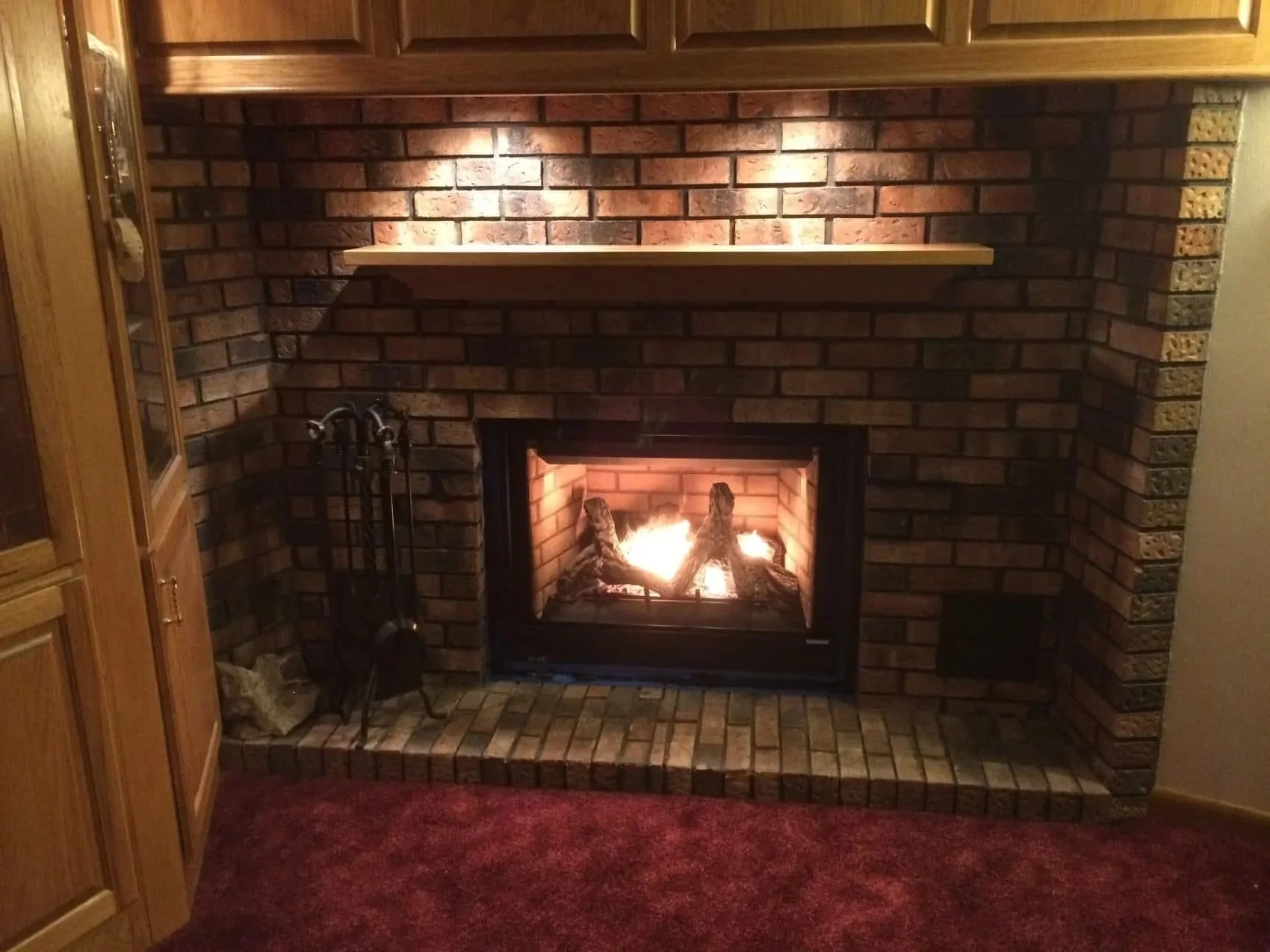 A cozy brick fireplace with a fire burning, a wooden mantel above it, and a set of fireplace tools to the side, in a living room with a carpeted floor.