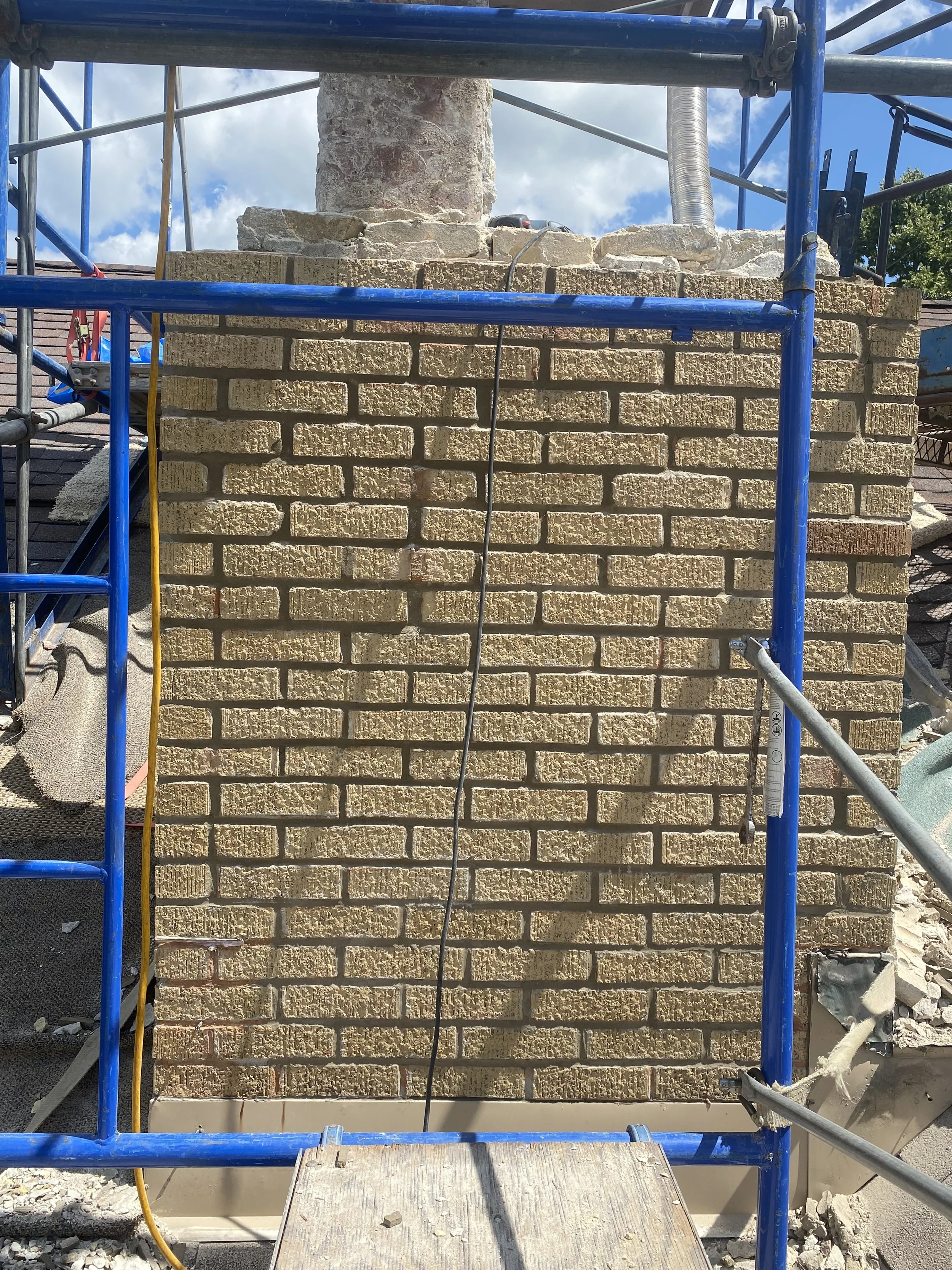 A brick chimney under construction surrounded by blue scaffolding, with a cloudy sky in the background.