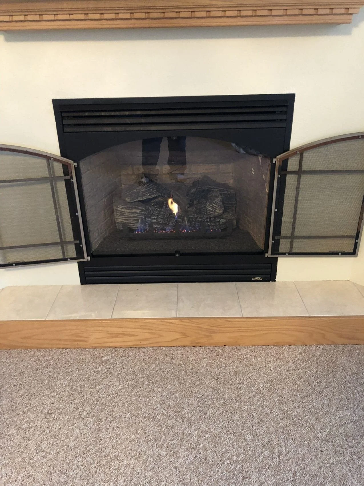 A fireplace with glass doors and a wood mantel, showing flames and logs inside, set on a tiled hearth with carpeted floor in front.