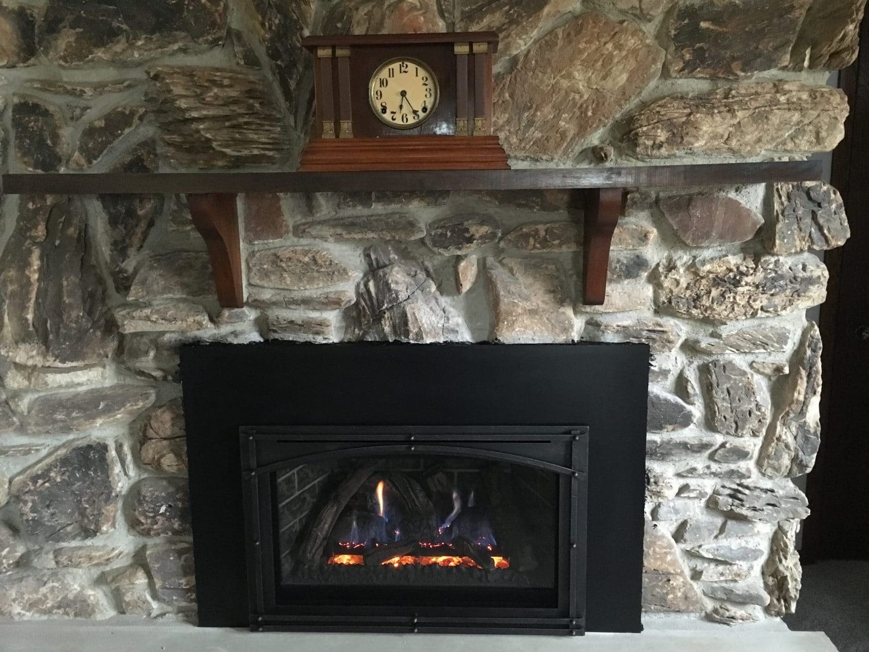 A fireplace made of large uneven stones with a black metal grate at the bottom. Above the fireplace is a wooden mantel shelf with a vintage wooden clock on it, set against a stone wall background.