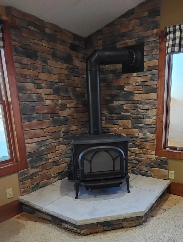 Wood stove in a corner with a stone wall background and a small tiled hearth, flanked by two windows with curtains.