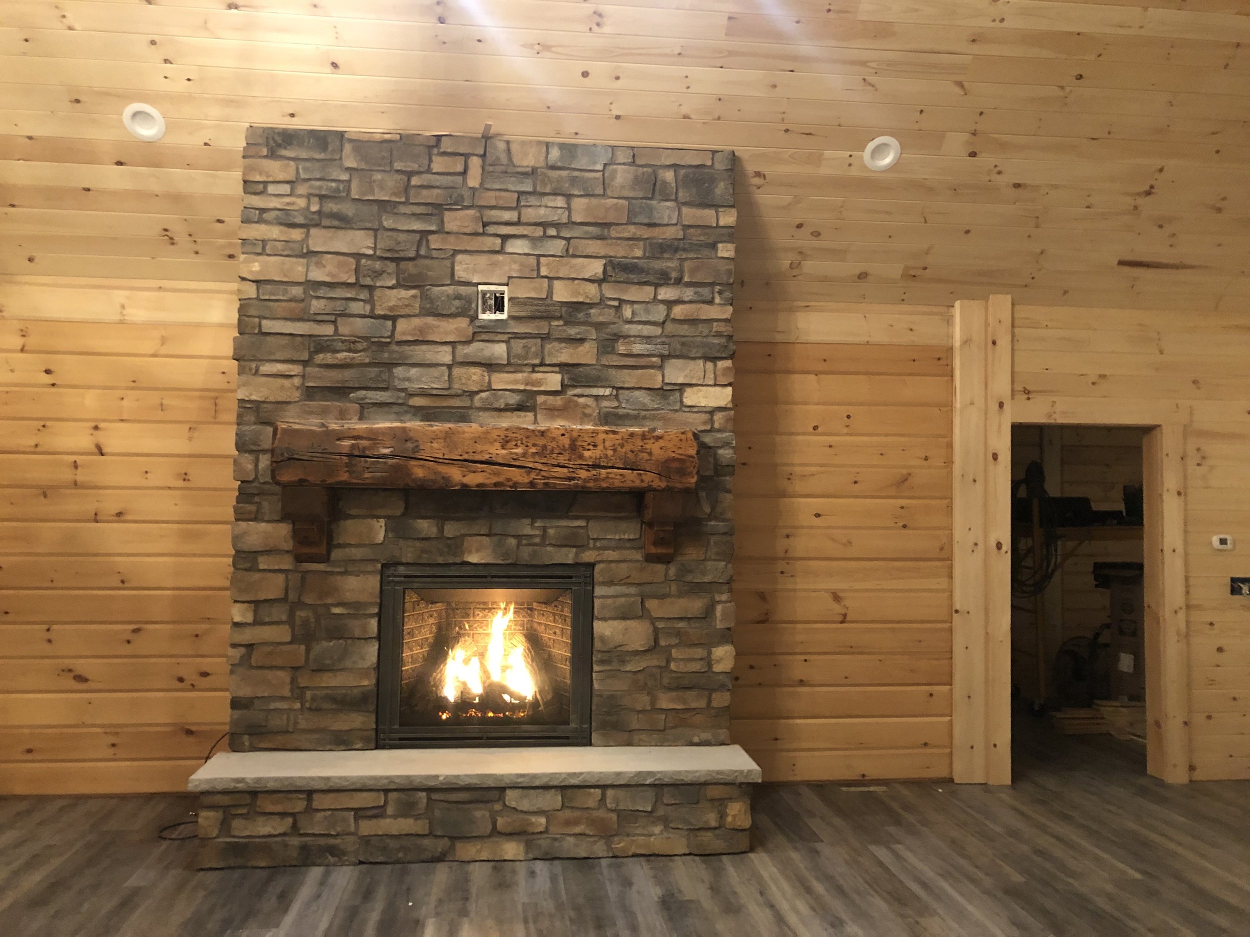 A rustic living room with wood-paneled walls and ceiling, featuring a stone fireplace with a wooden mantel and a fire burning inside.