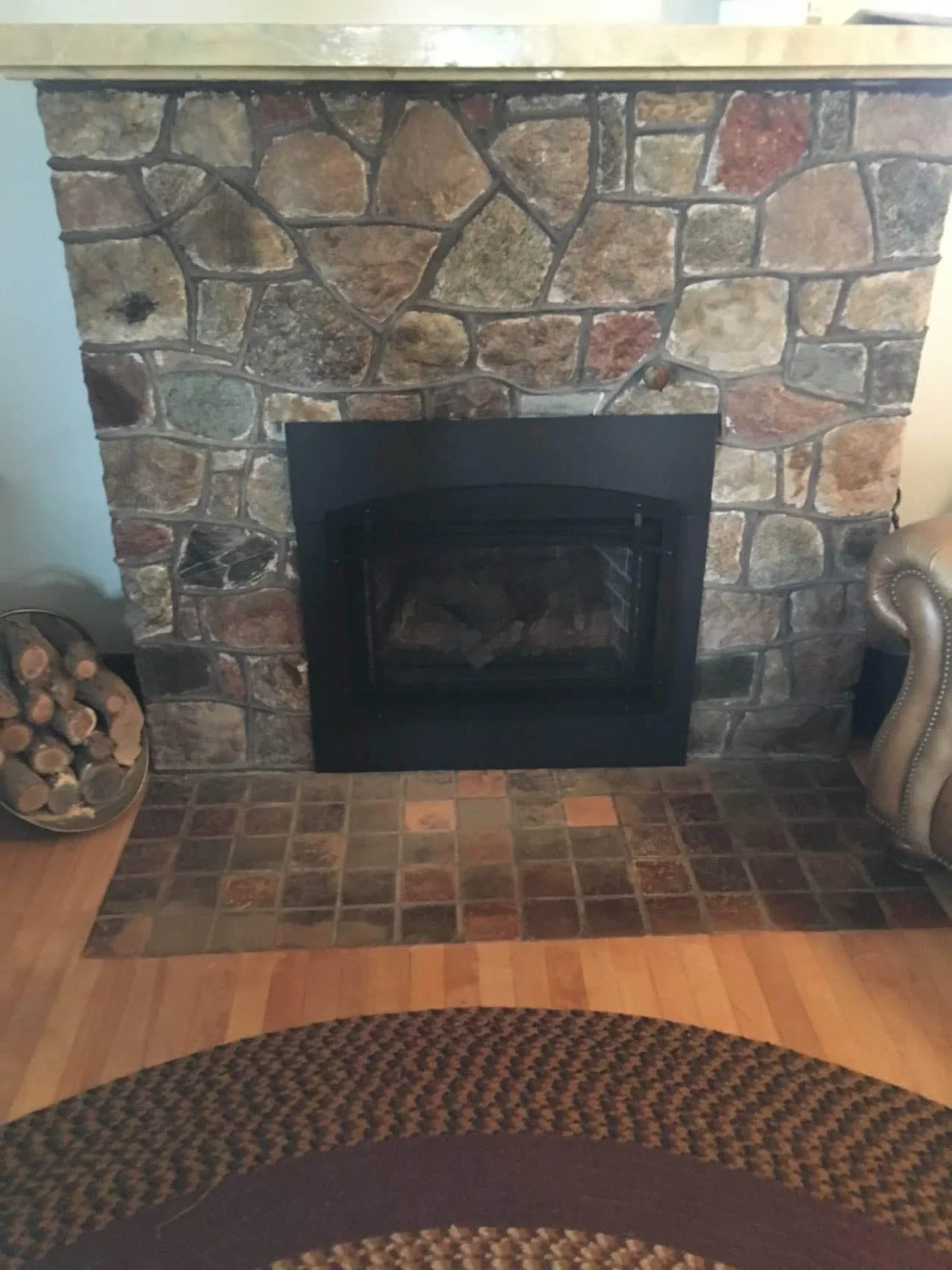 Stone fireplace with a wood stove insert, a wood log holder on the left, and part of a brown patterned rug in the foreground.
