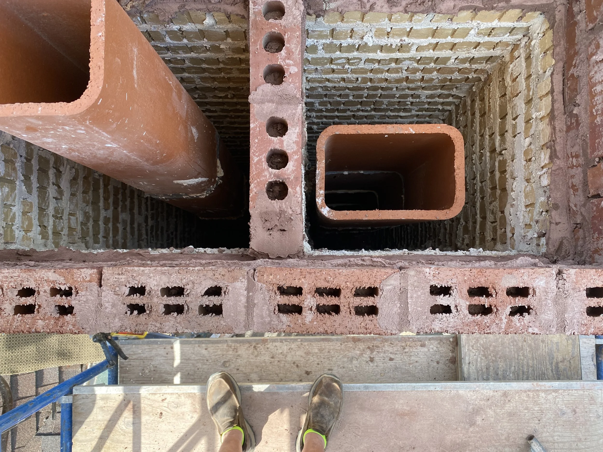 View from a construction scaffold showing a person wearing yellow-green sneakers on wooden planks, looking down at a brick structure with open square and circular holes, in a building under construction.