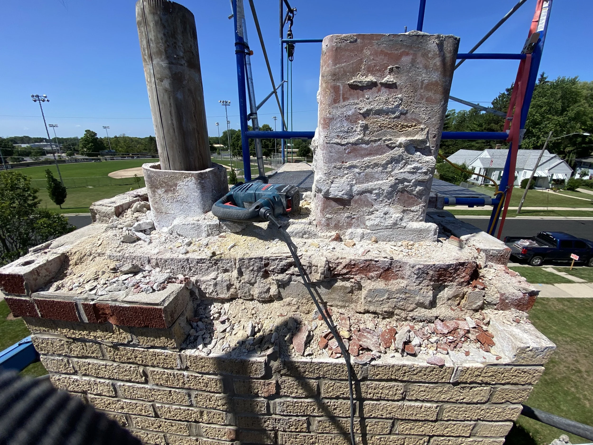 Construction site with a partially built brick and stone wall, scaffolding, and various construction tools including a power drill, against a background of a baseball field and blue sky.