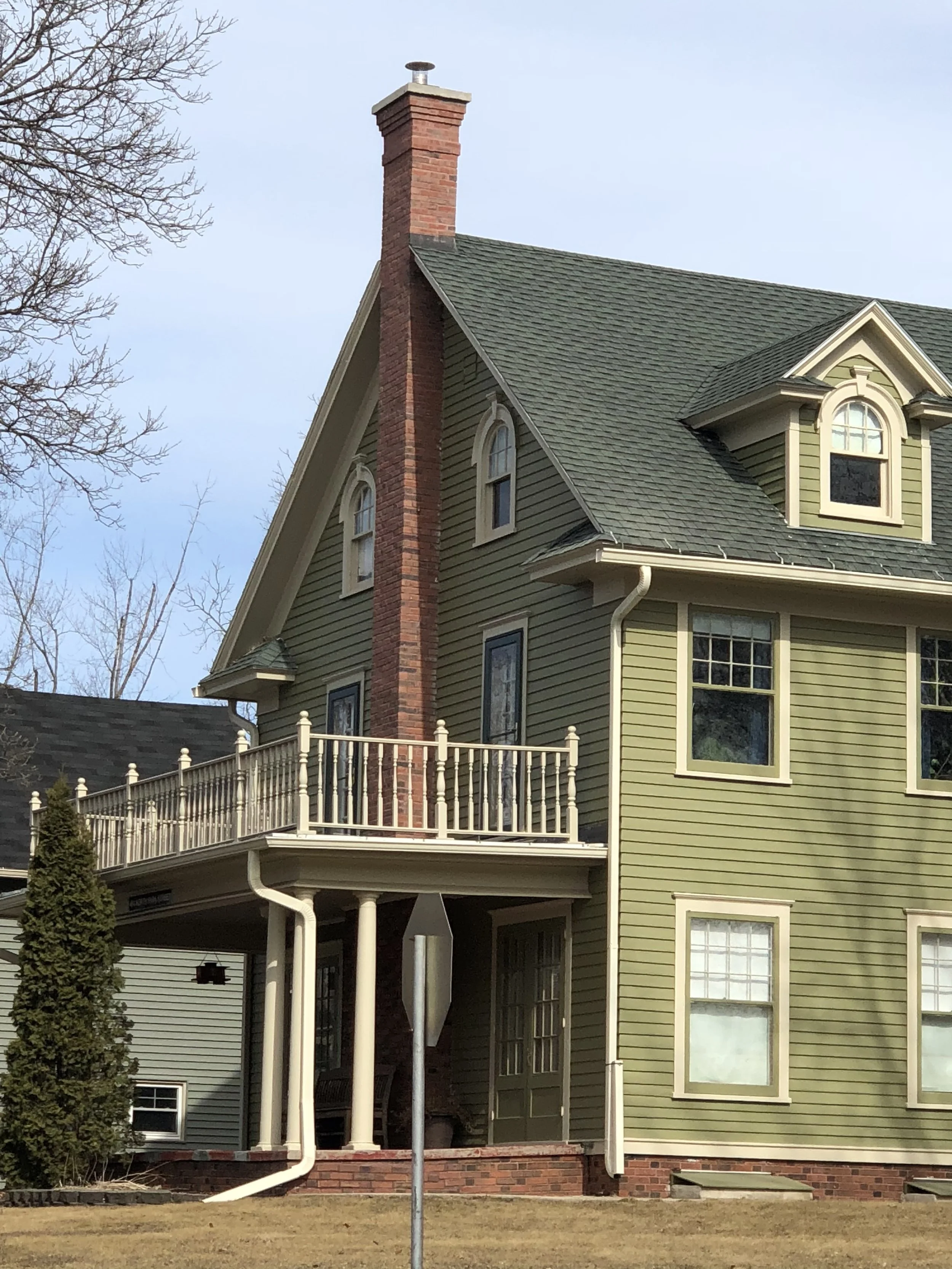 A two-story house with green siding, multiple windows, a brick chimney, a balcony with white railing, and a backyard with a tree and dry grass.