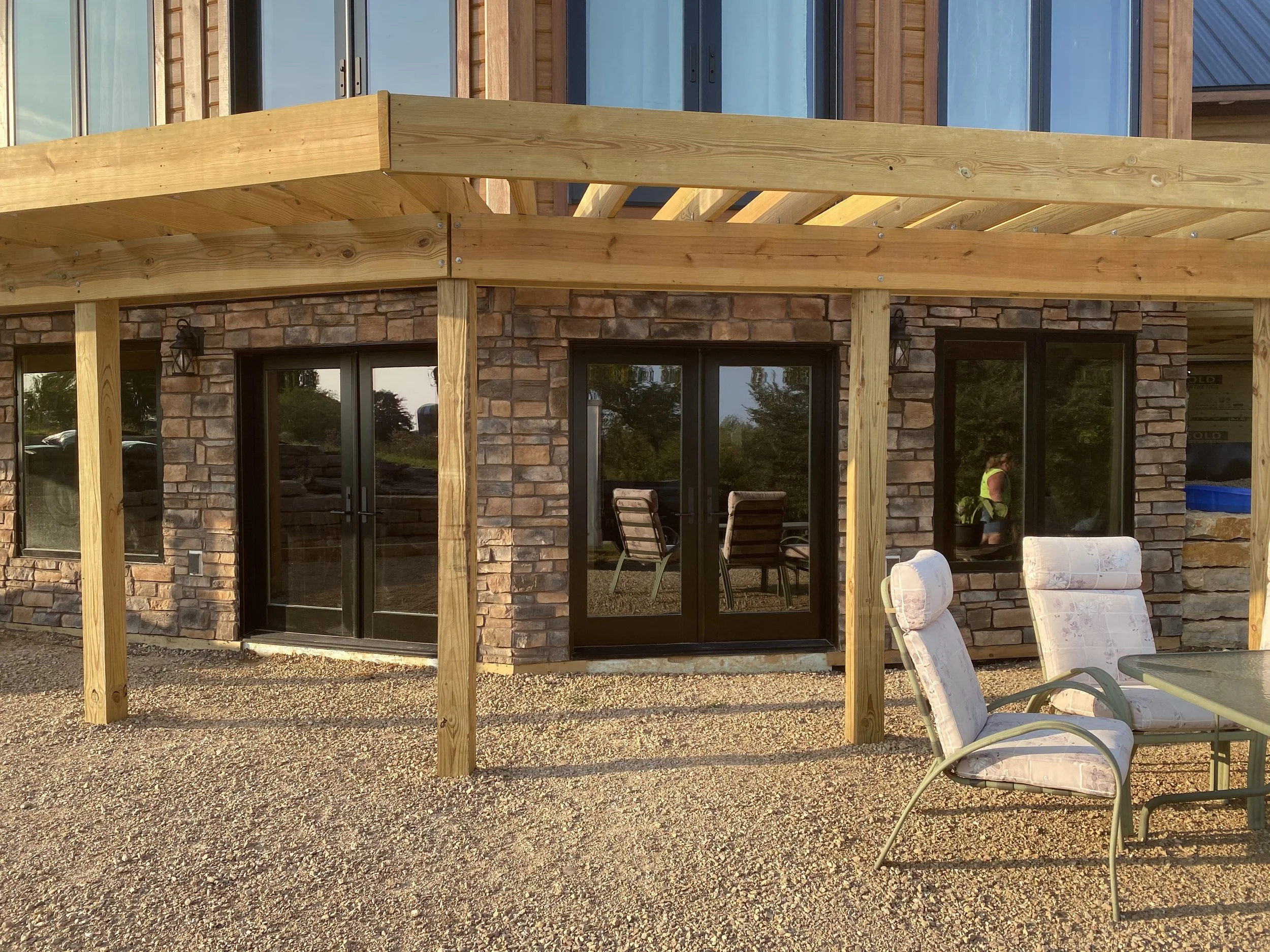 A new wooden deck under construction in front of a house with brick walls and sliding glass doors. Patio chairs and a person in a vest are visible in the yard.