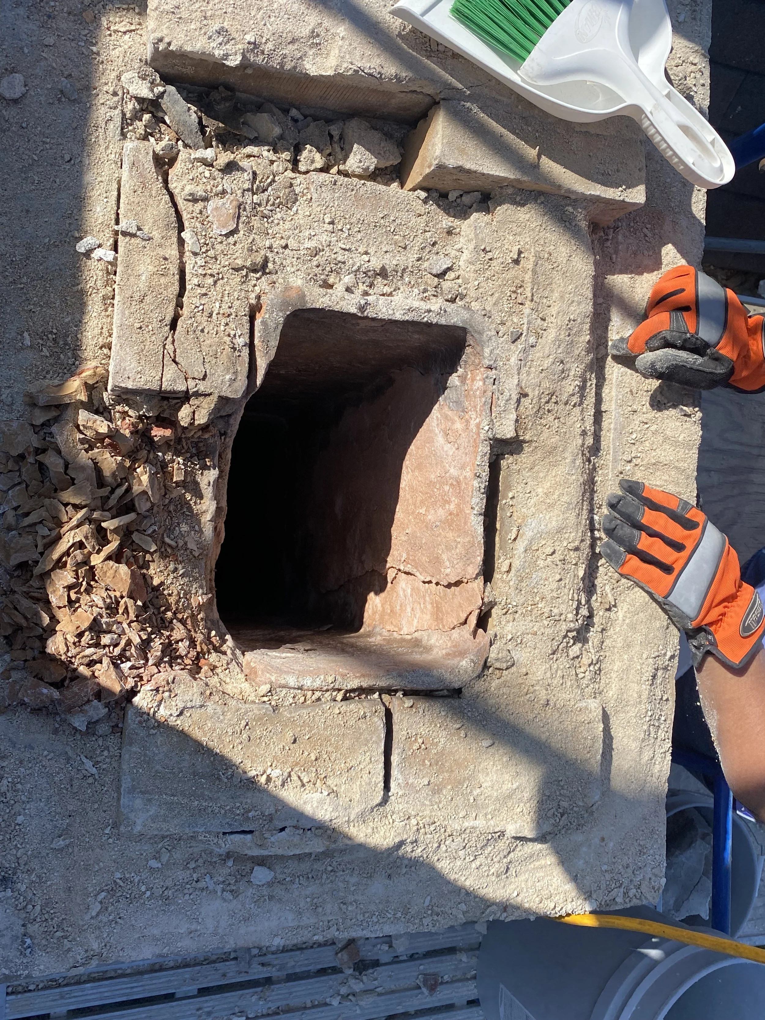 A brick chimney opening under repair in a construction site, with tools such as gloves and a trowel nearby, and construction materials like bricks and debris around.