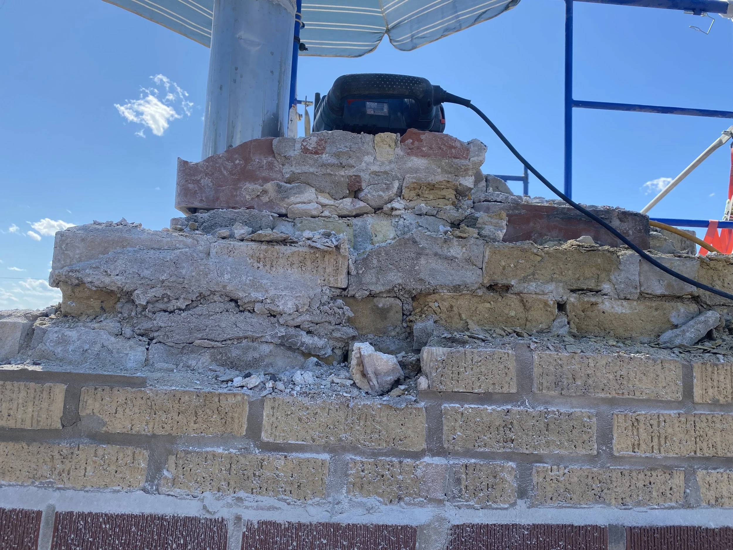 Close-up of a construction site with weathered bricks and stones, a Bosch electric tool connected by a black cord, scaffolding and a blue sky in the background.