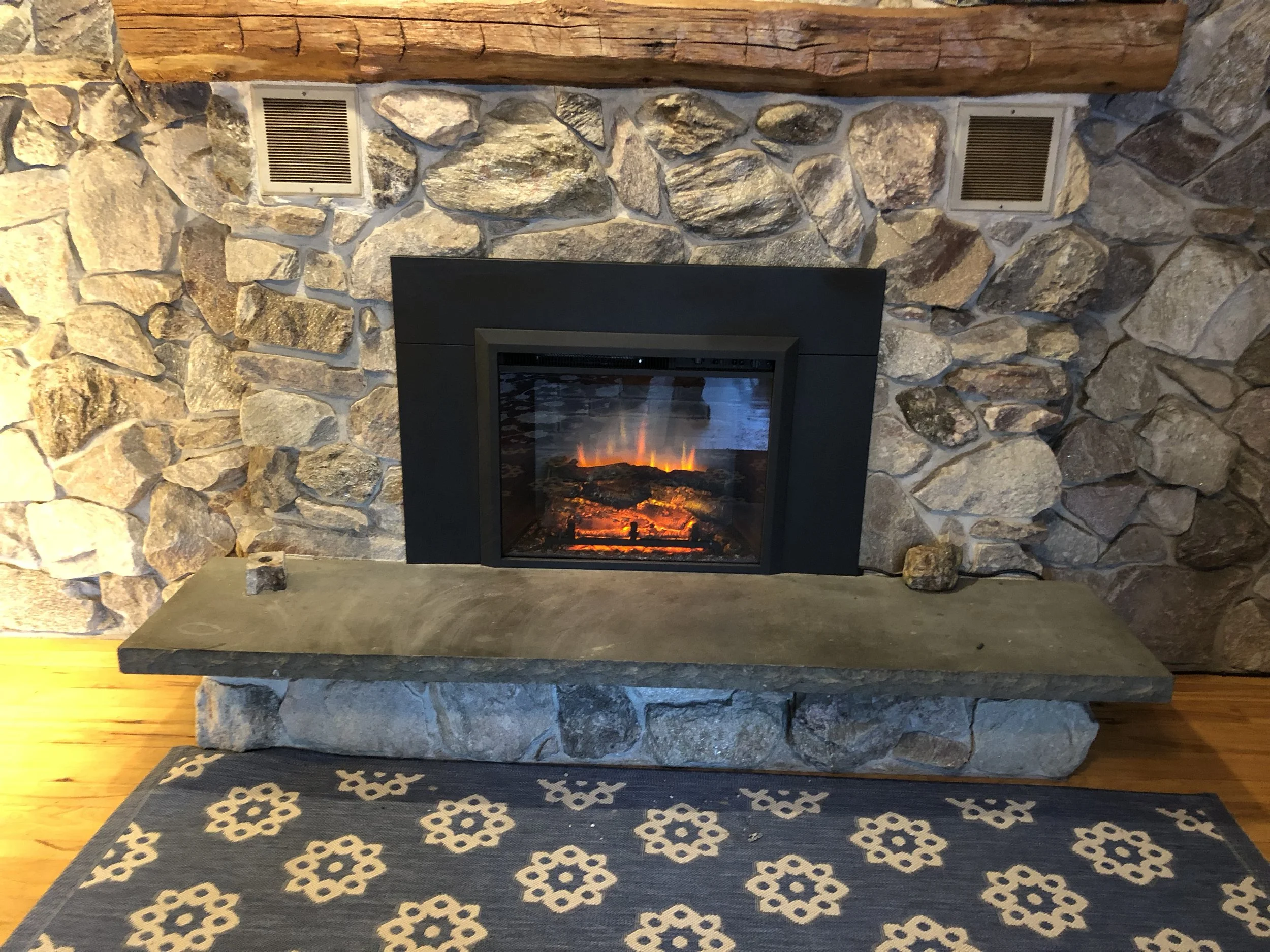 A stone fireplace with two vents near the top and a wooden mantel. There is a cozy rug with a floral pattern in front of the fireplace, and a piece of wood on the bottom right corner of the fireplace.