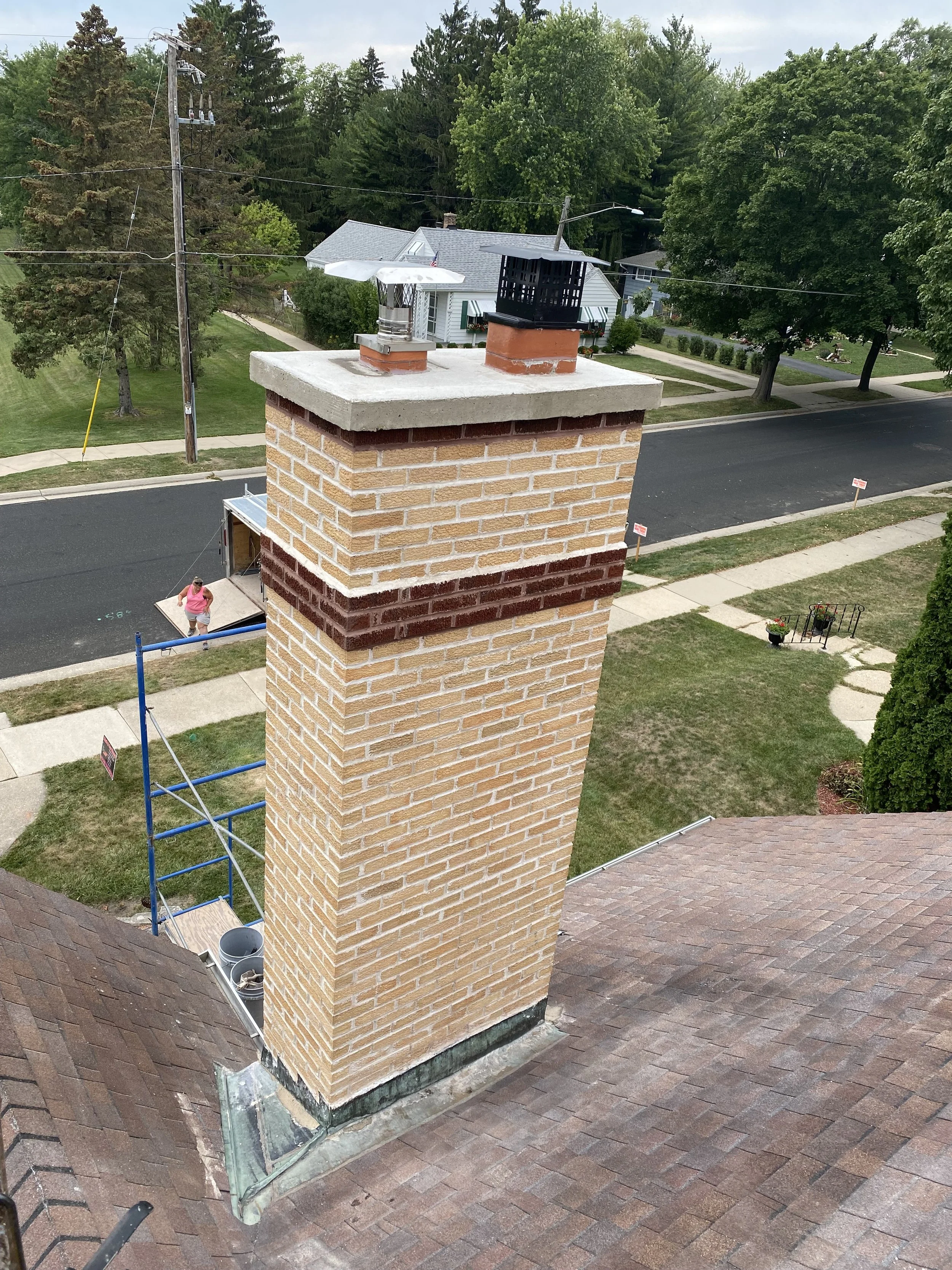 A large brick chimney extends above the roof of a house, with two smaller chimney structures on top, one with a white cap and the other with a black cover. In the background, there are trees and a street with houses, and a person is walking on the si