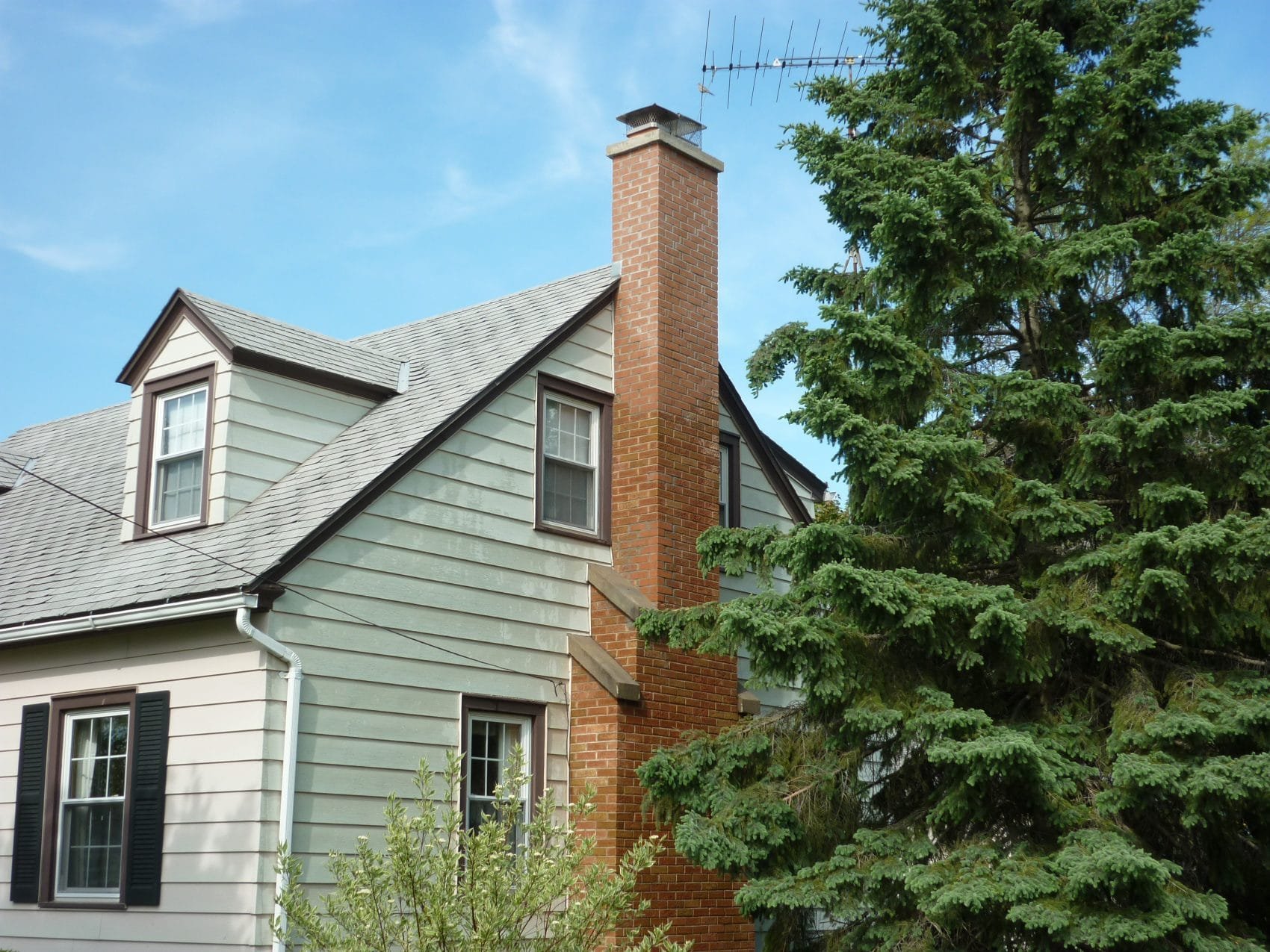 Photo of a two-story house with gray siding, black window shutters, a brick chimney, and an evergreen tree nearby under a blue sky.