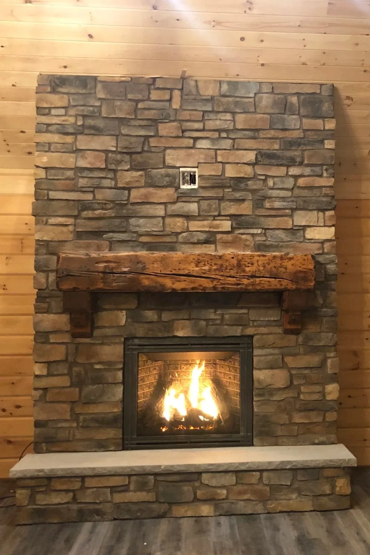A stone fireplace with a wooden mantel and a fire burning inside adorned with a wood-paneled wall behind it.