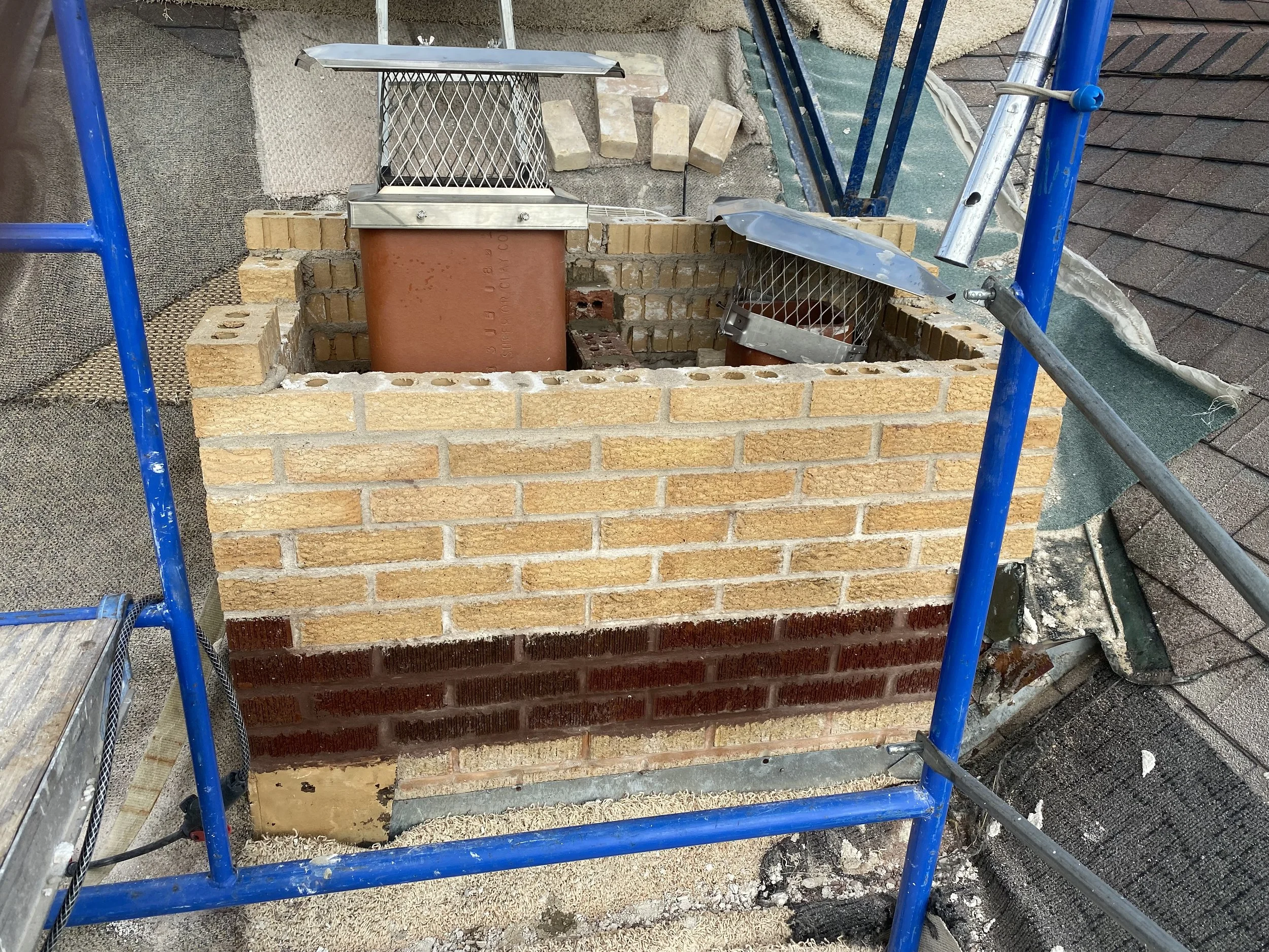 A brick fireplace under construction with a metal chimney pipe surrounded by yellow and red bricks. There is blue scaffolding on the sides and construction materials around.