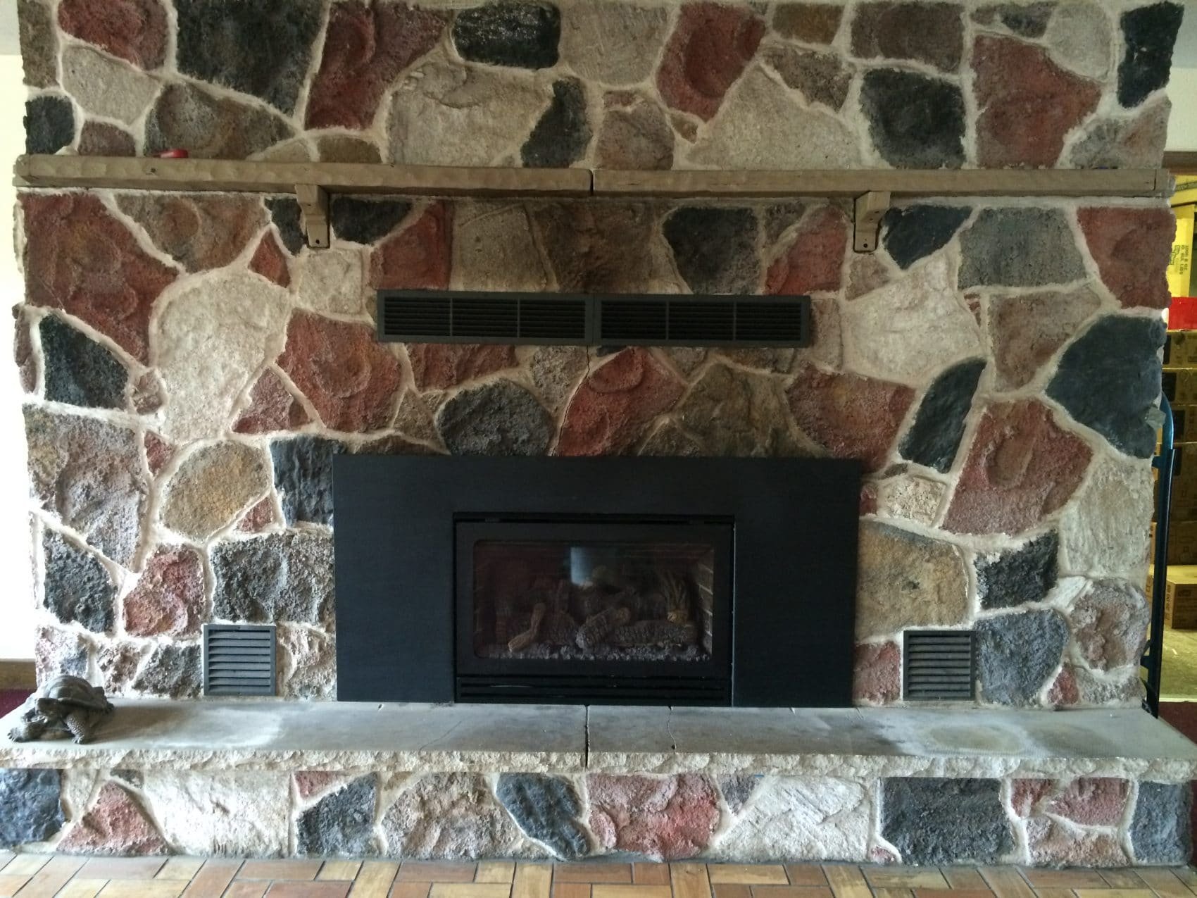 A stone fireplace with a black metal insert, a wooden mantel, and a decorative lion figurine on the stone hearth. The stones are irregularly shaped and multicolored, with shades of beige, red, black, and gray.