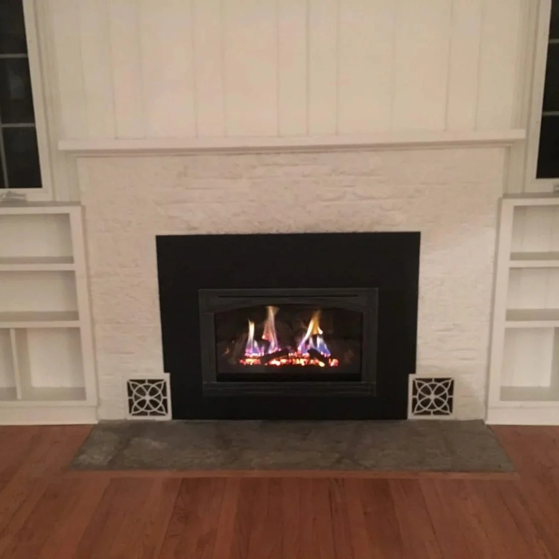 Fireplace with flames burning inside, surrounded by white built-in shelves and a wooden floor.
