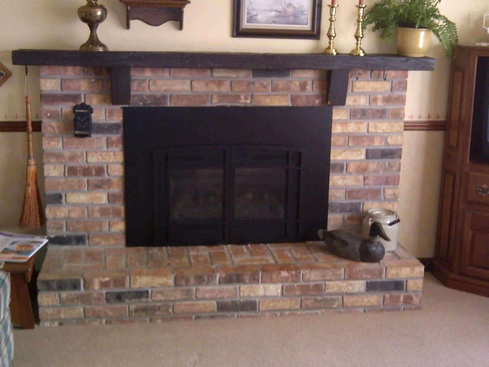 A brick fireplace with a dark wooden mantel, decorated with brass candlesticks, a decorative urn, and a potted fern plant. There is a framed painting above the mantel and a small lantern hanging on the left side of the fireplace. On the right side, a