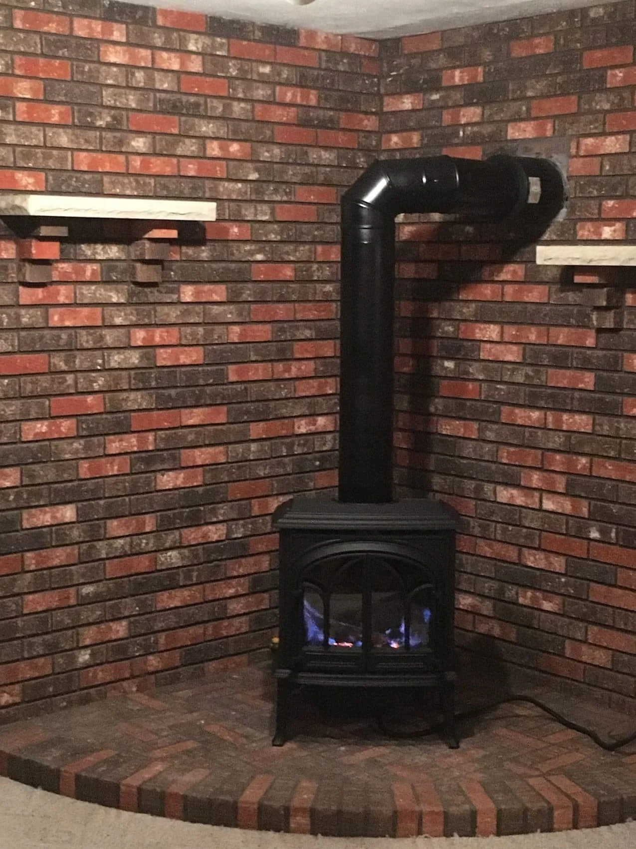 Brick fireplace with a black wood stove and a black stovepipe against a brick wall with two small wall-mounted shelves.