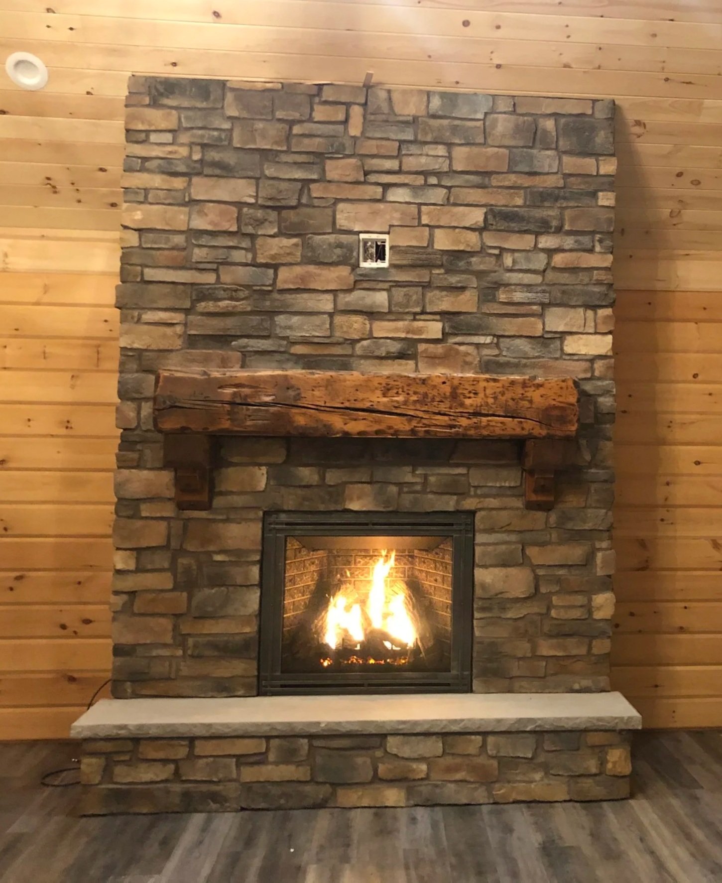 A stone fireplace with a wooden mantel in a room with wood-paneled walls and hardwood flooring, with a fire burning inside.