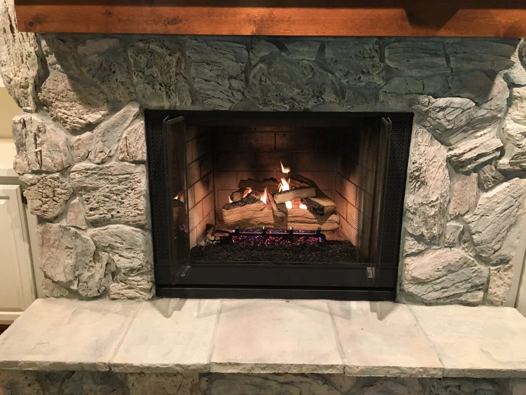 A stone fireplace with a wooden mantle, containing a small fire with logs and glowing embers inside.