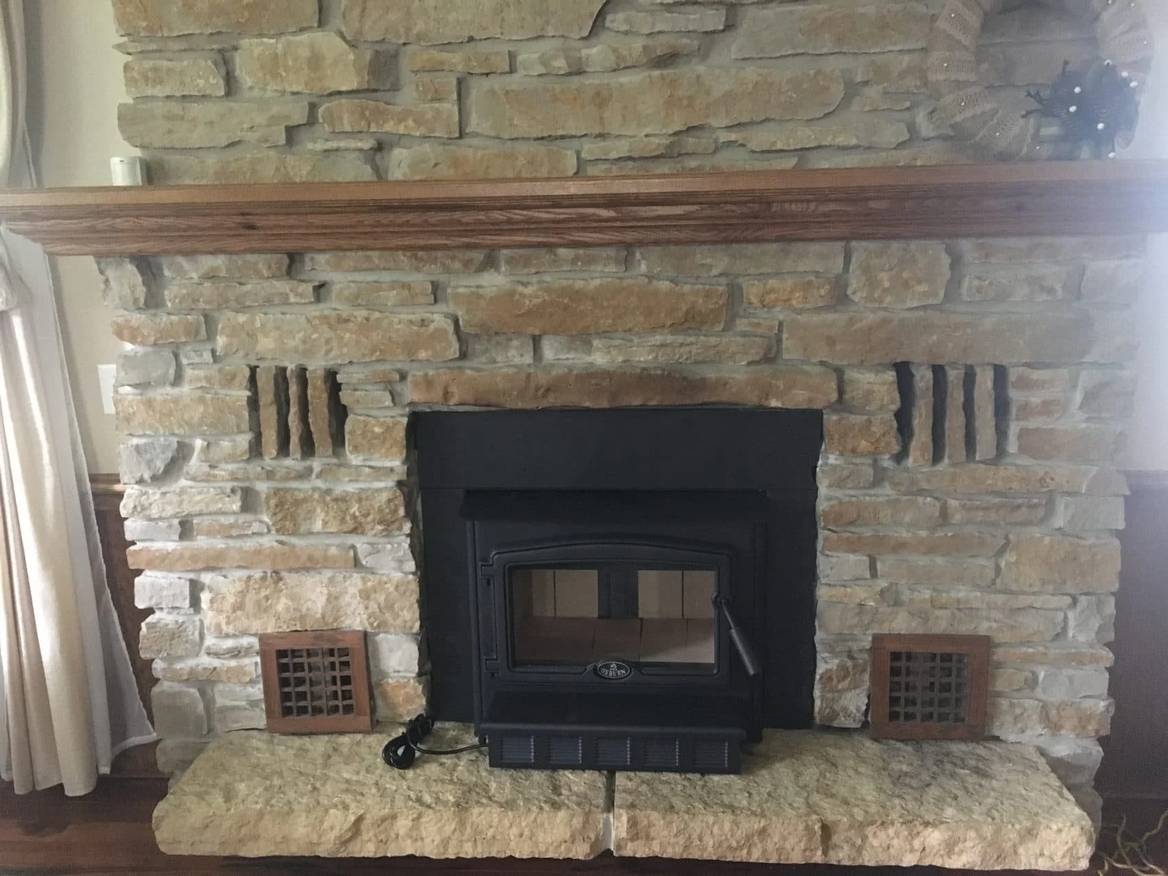 Stone fireplace with a black wood stove insert, wooden mantel, and two small side vent panels.