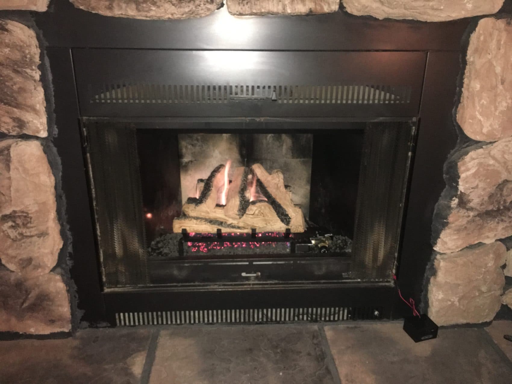 A fireplace with a stone surround and a metal frame, with logs burning inside and glowing embers, in a cozy indoor setting.