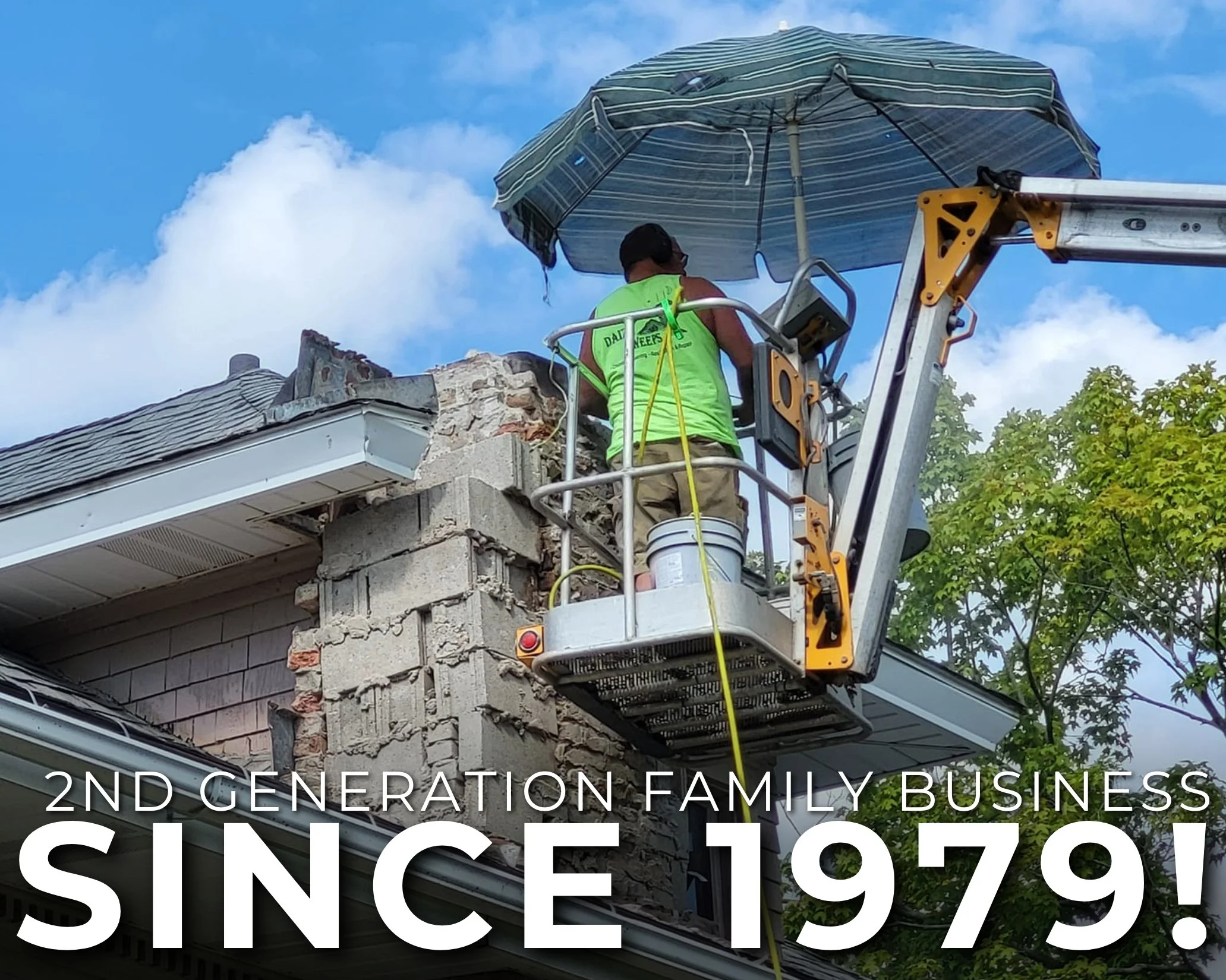 A construction worker on a lift working on the exterior of a two-story house with damaged siding and exposed brick. The worker is wearing a neon green vest and a black cap, and there is a large striped patio umbrella above him. The sky is blue with some clouds, and there is green foliage in the background.