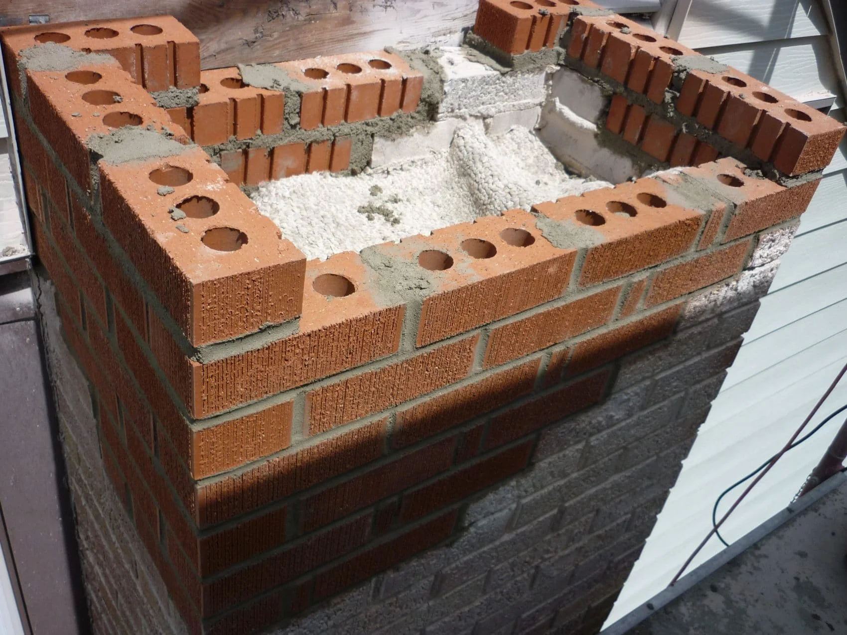 Close-up of a brick chimney under construction with red bricks, gray mortar, and some white insulating or filling material visible inside.