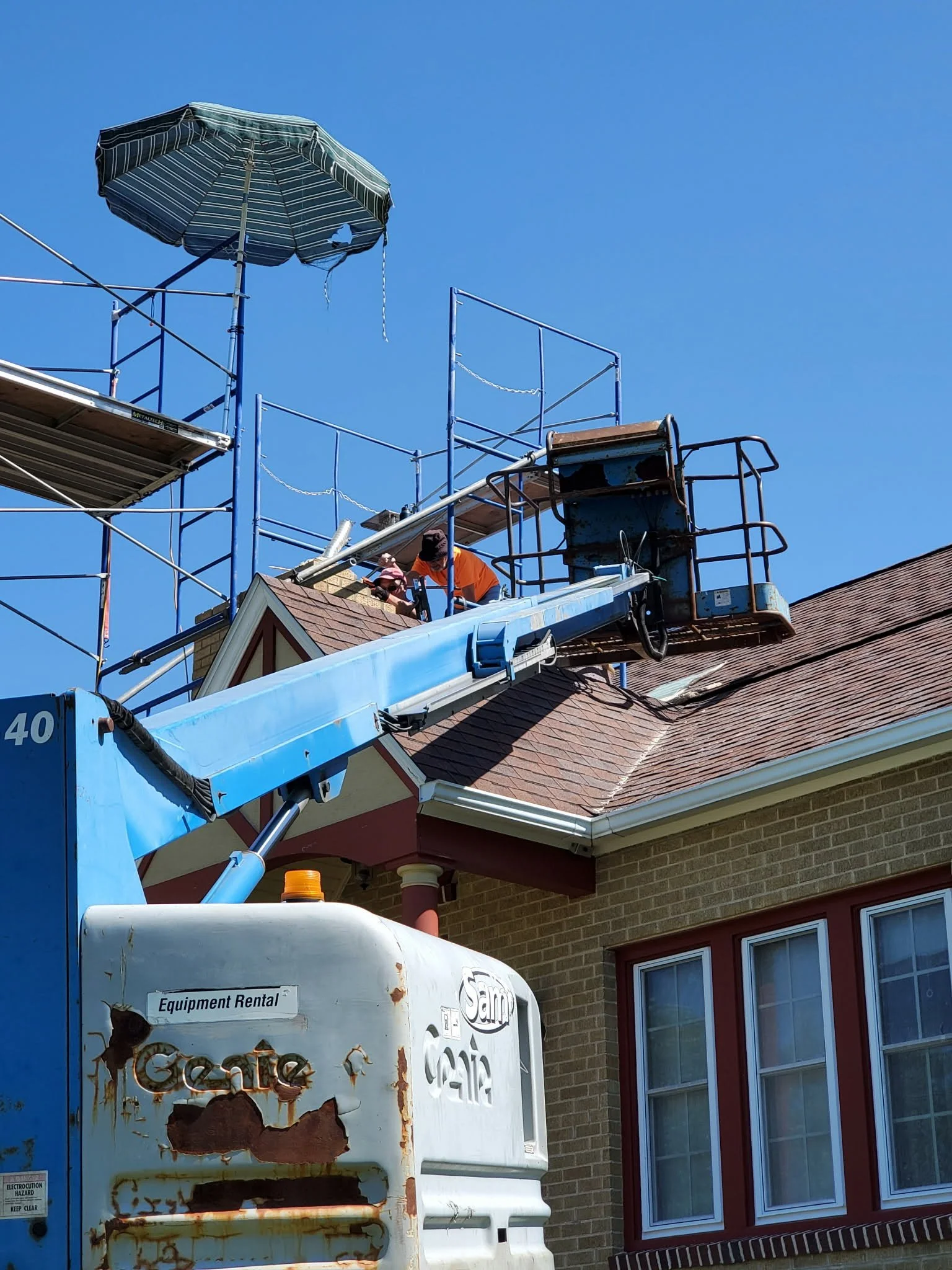 Workers on a roof near a rusted and damaged white equipment box, with a blue sky background and a house with brick exterior and red-framed windows.
