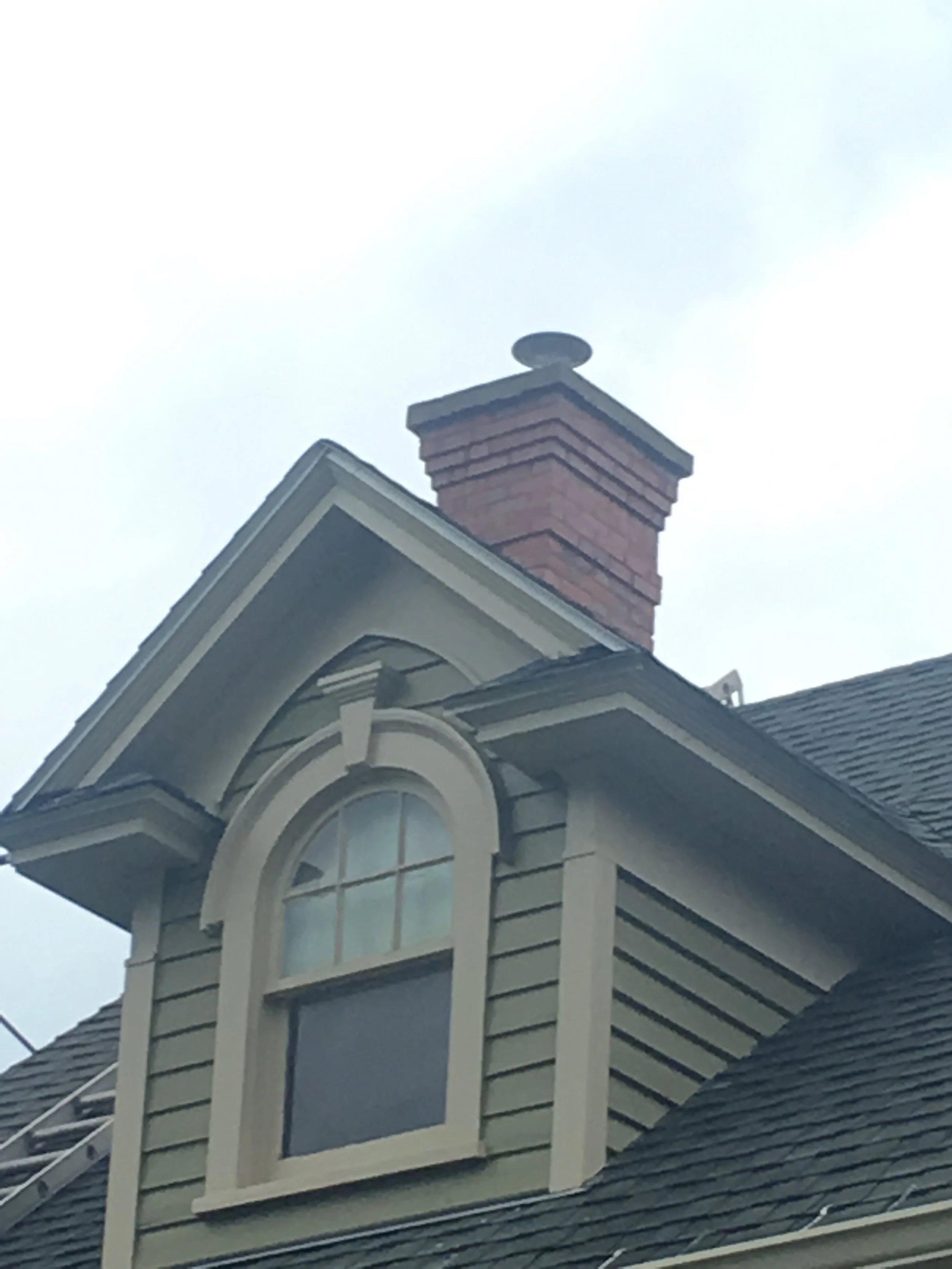 Close-up of a house dormer window with detailed wood trim, a arched window, and a red brick chimney on a residential roof.