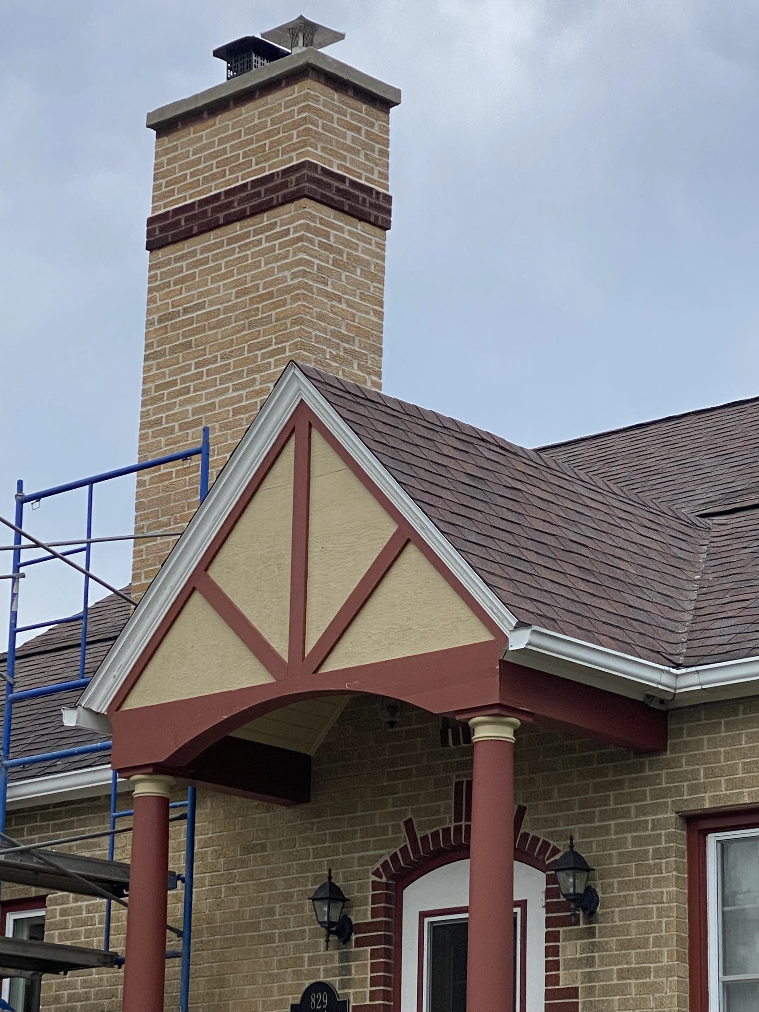 Close-up of a brick house with a chimney, covered porch with decorative trim, red columns, and black lantern-style porch lights. Part of construction scaffold is visible on the left.