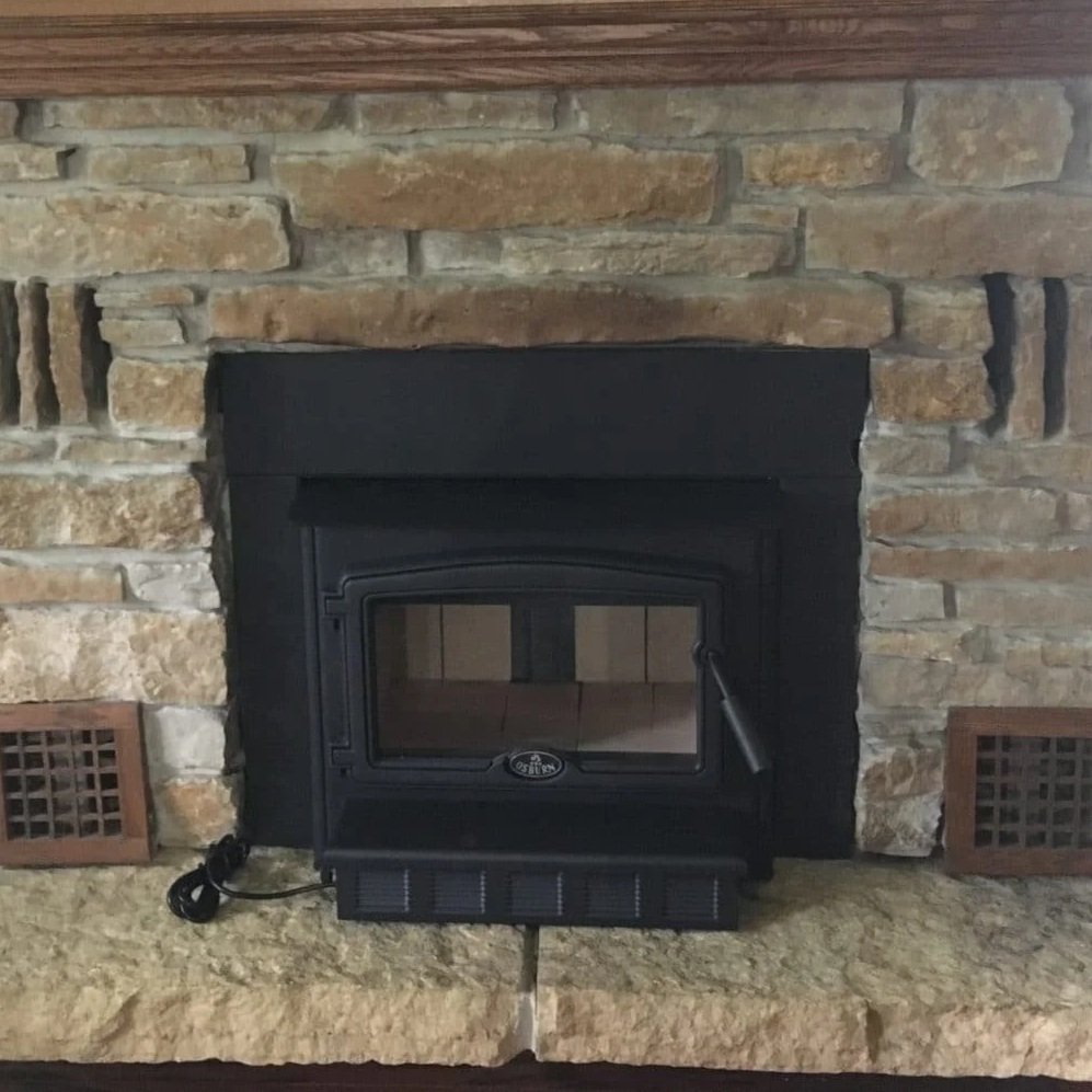 Black wood stove in front of a brick fireplace with a stone hearth and two air vents on each side.